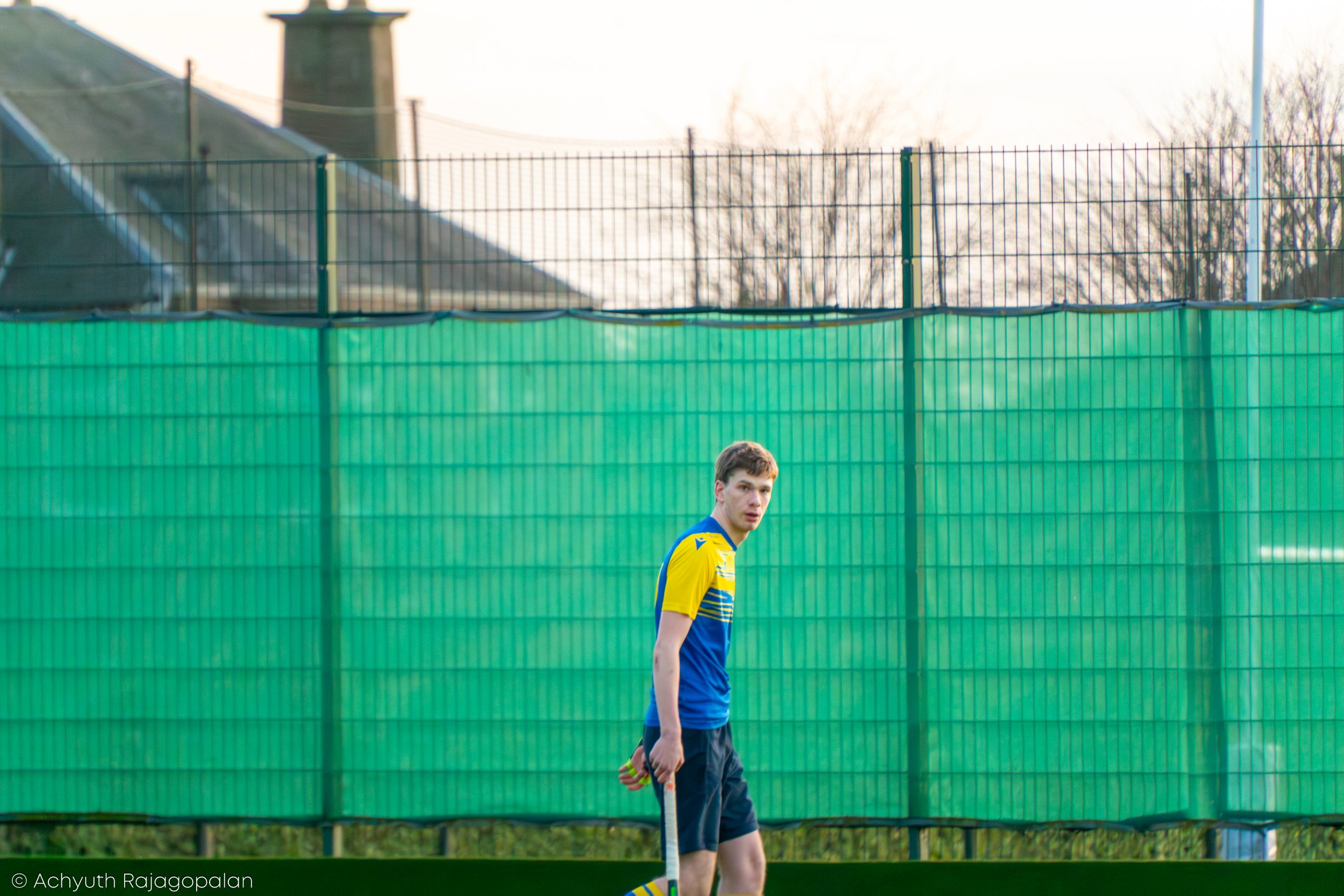A young man dressed in sportswear holding a hockey stick standing in front of a green fence on a sports field, with trees and a building in the background.