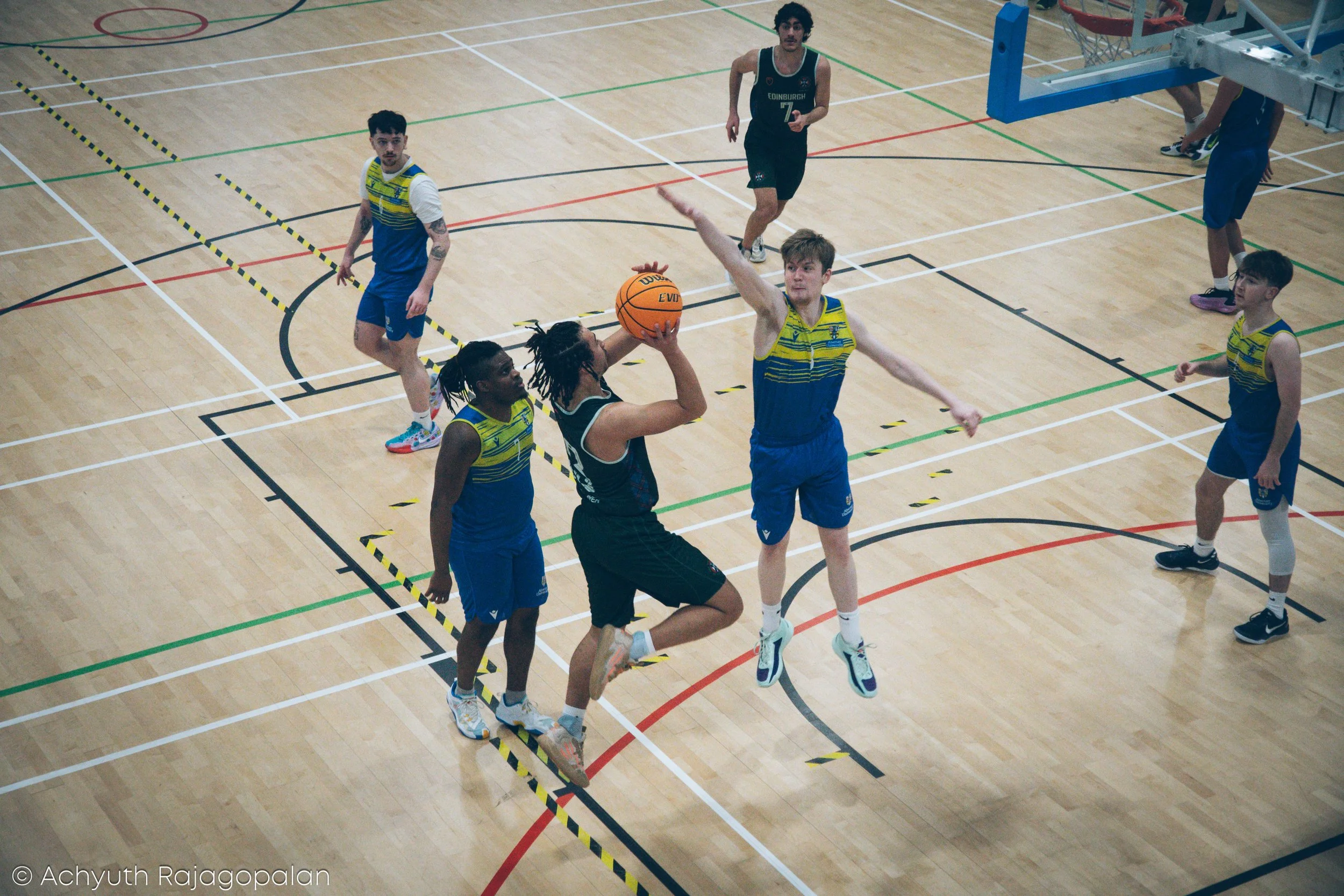 A group of young men playing basketball indoors on a basketball court, with some players jumping and reaching for the ball near the basket.