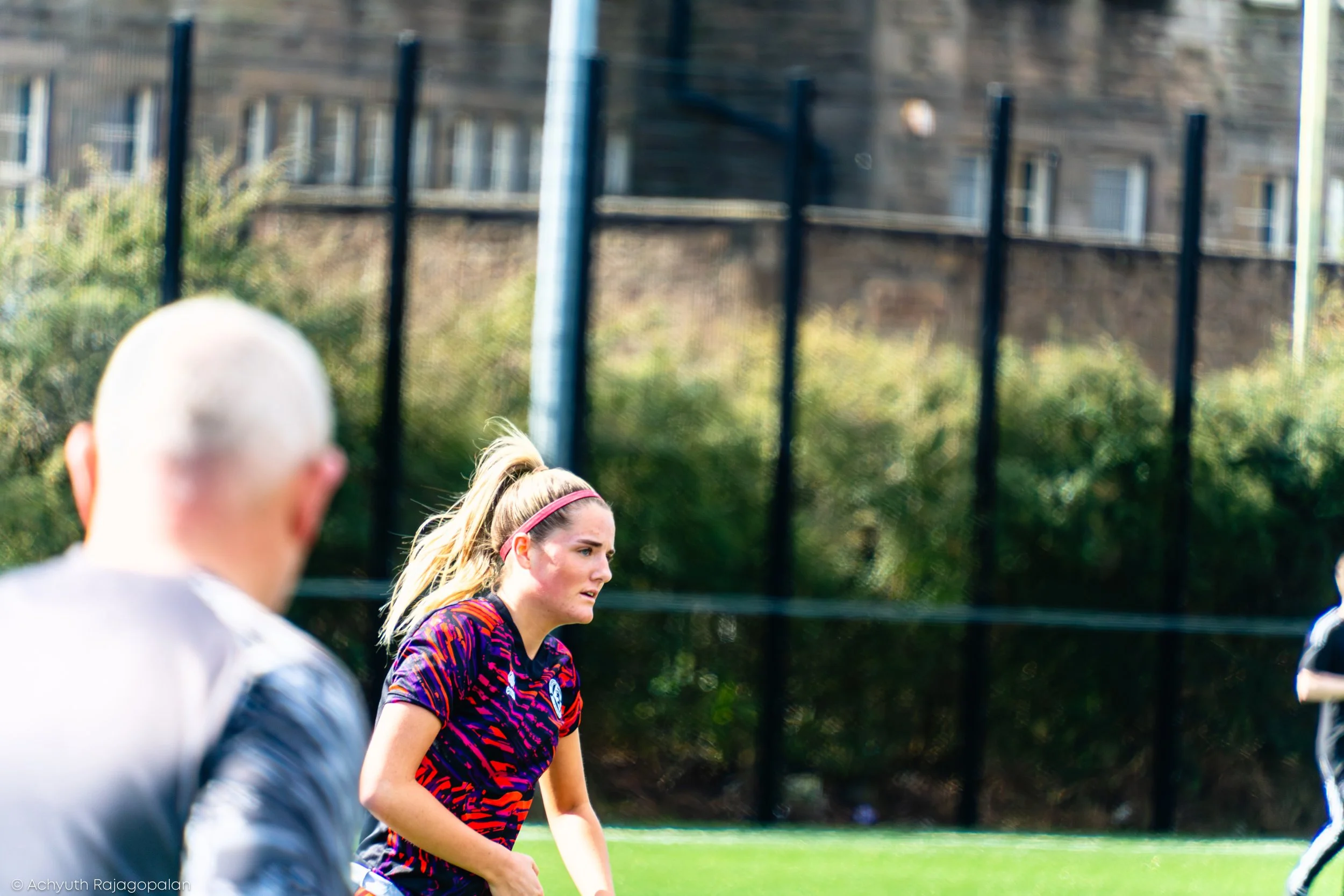 A woman in a colorful sports shirt running outdoors with a group of people, with a fence and a building in the background.