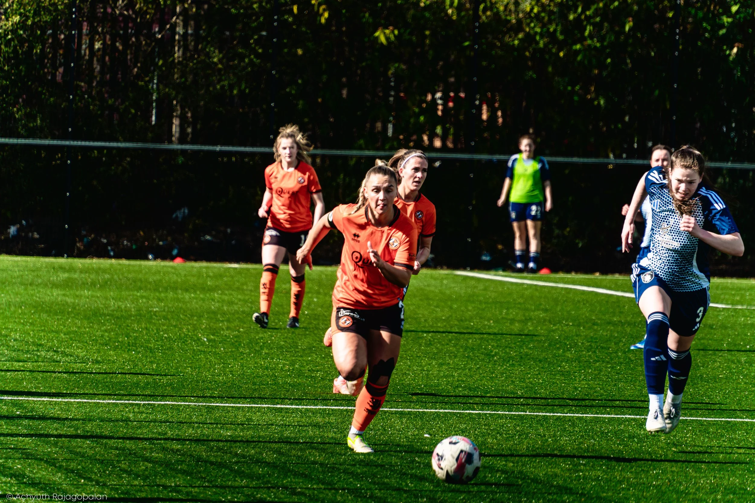 A group of women soccer players running on a field during a game, with some players wearing orange jerseys and others in blue jerseys. One player in the foreground is about to kick a soccer ball.