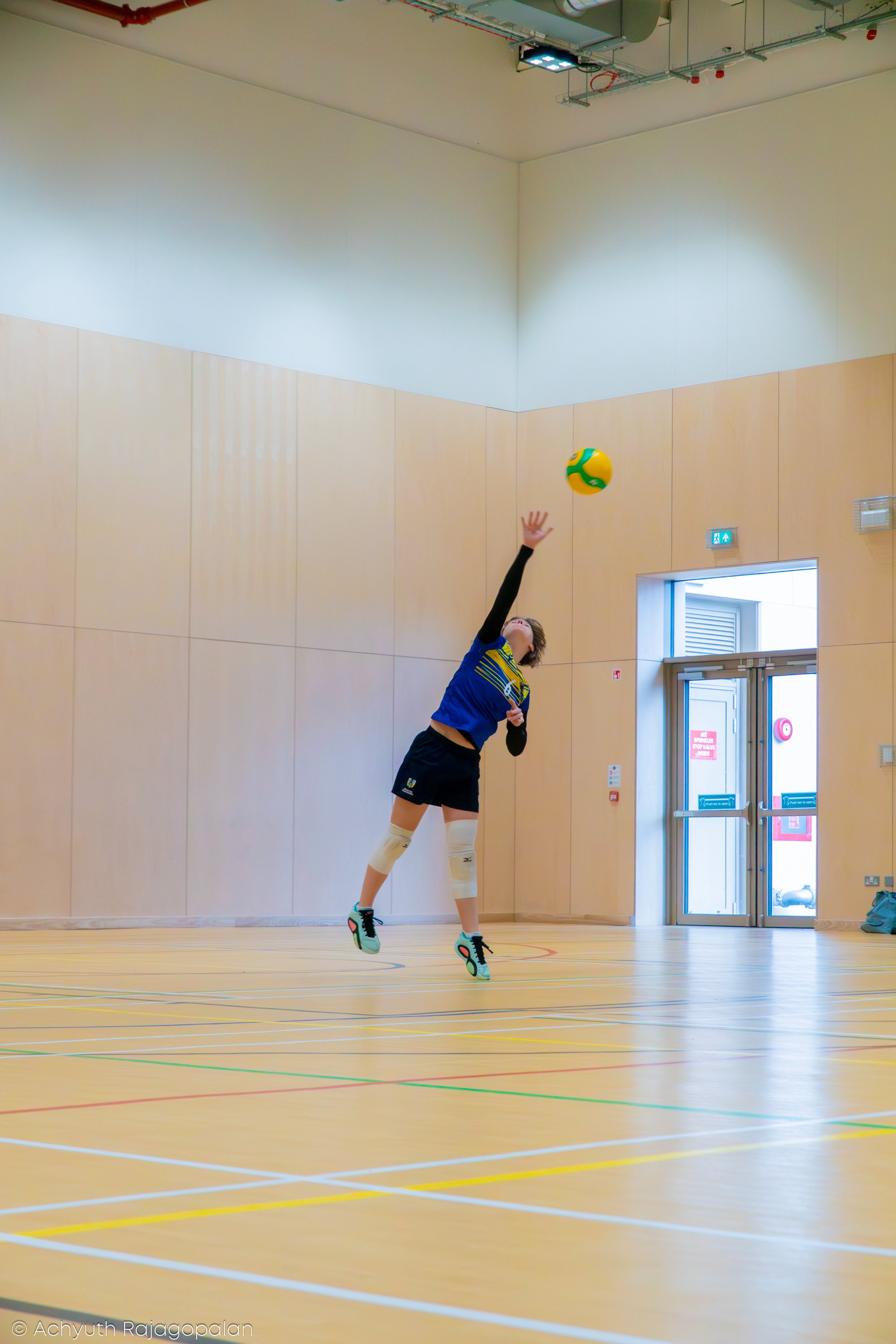 A person playing volleyball indoors, jumping to hit a yellow and green volleyball. The person has knee-high white socks, athletic shoes, and is wearing a blue and yellow athletic shirt. The indoor gym has a wooden floor and beige wall panels, with an