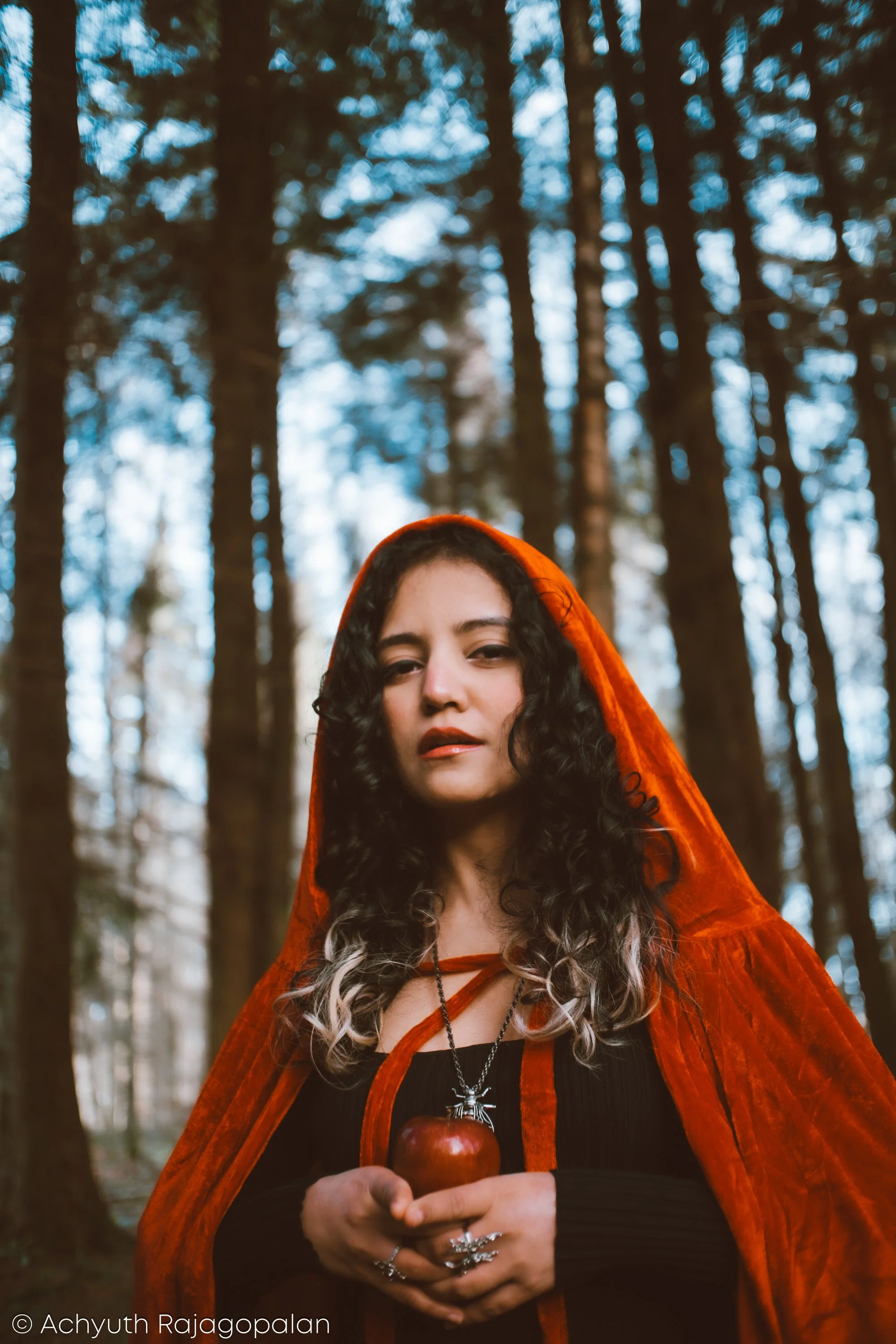 A woman with curly dark hair, wearing a red hooded cloak, standing in a forest of tall trees with blue sky visible through the branches, holding a red apple in her hands.