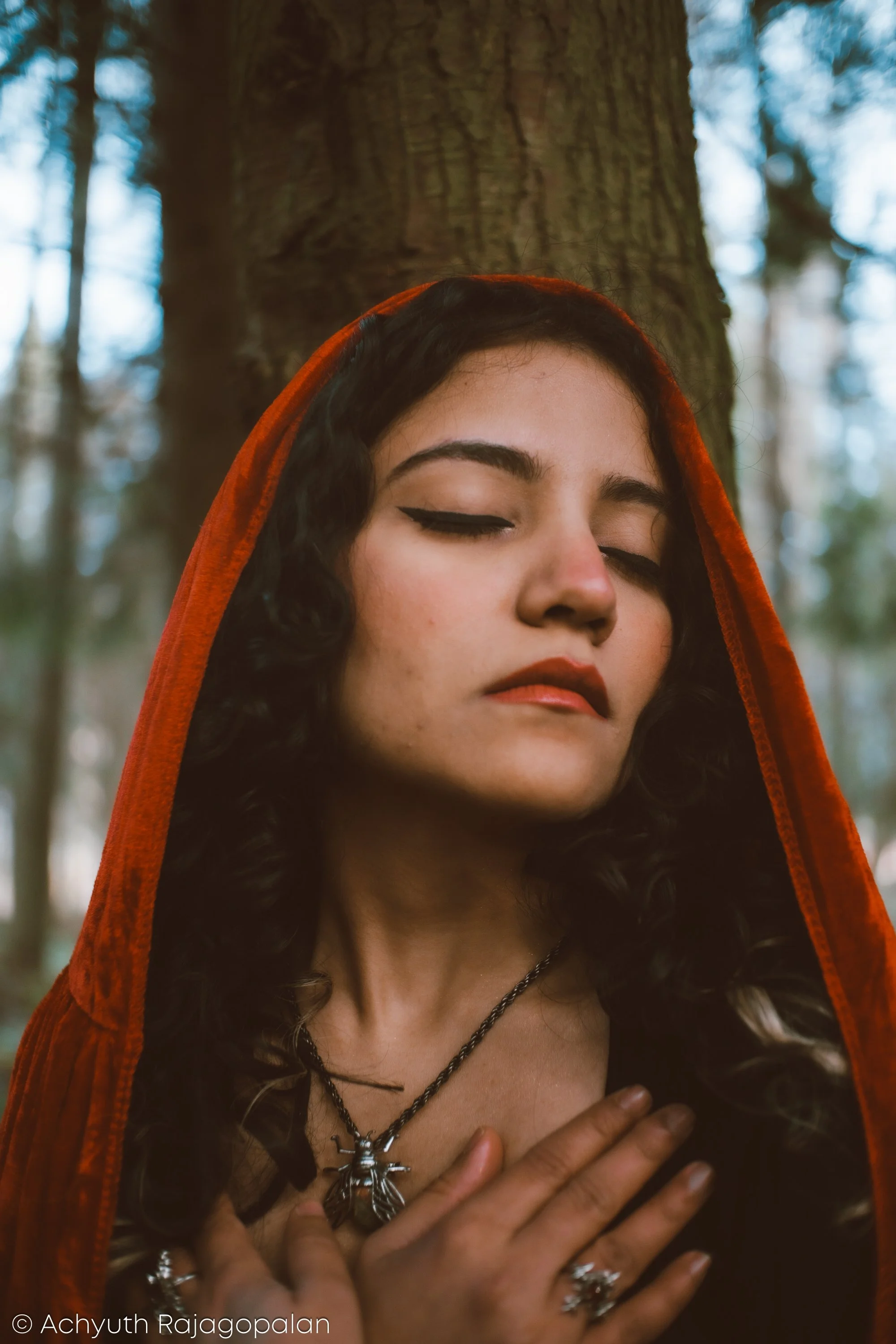 A woman with dark curly hair wearing a red hooded cloak and ornate jewelry, including a cross necklace and rings, standing in a forest with her eyes closed and hand on her chest.
