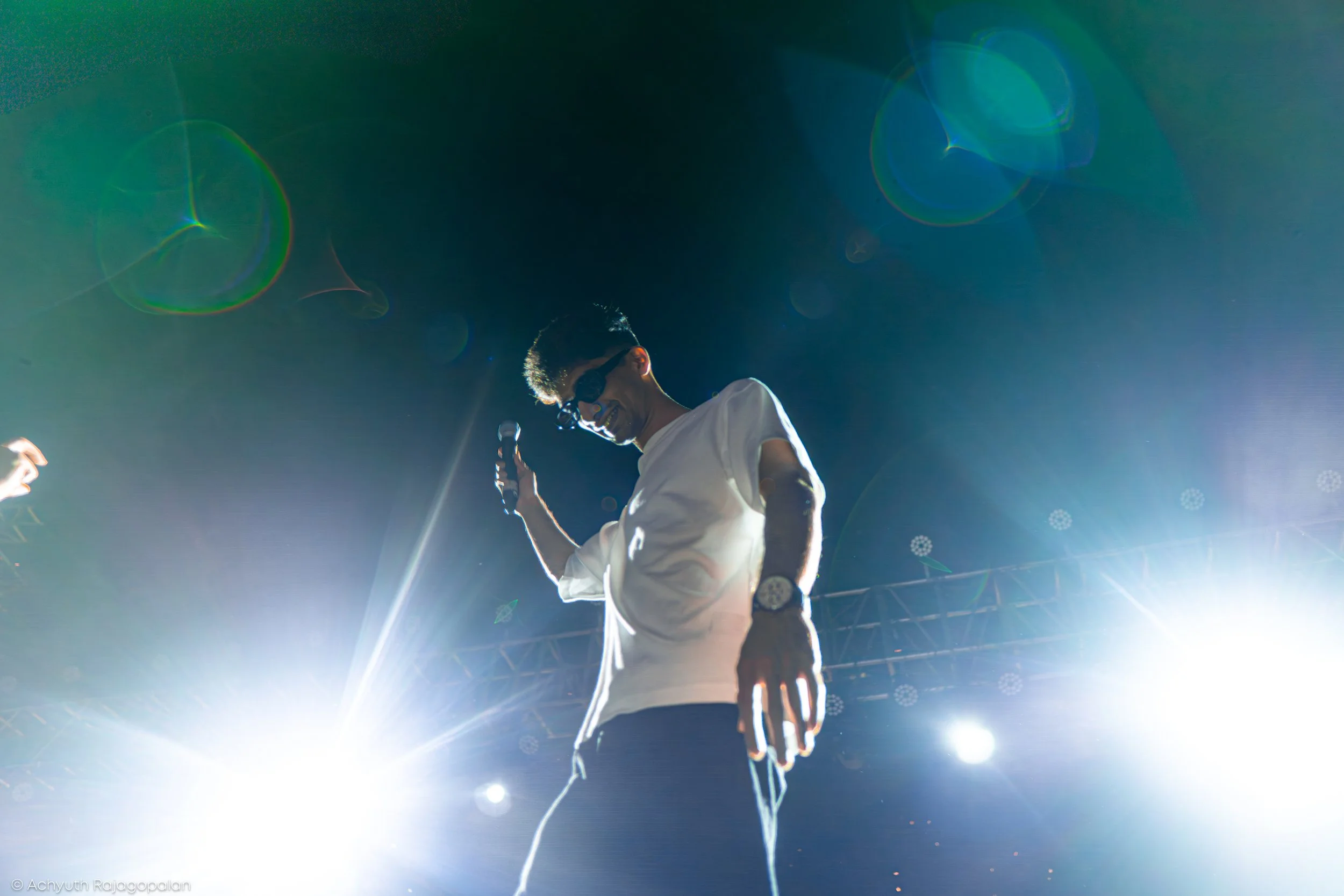 A man wearing sunglasses and a white shirt holding a microphone, illuminated by bright stage lights, is performing on stage with a dark background.