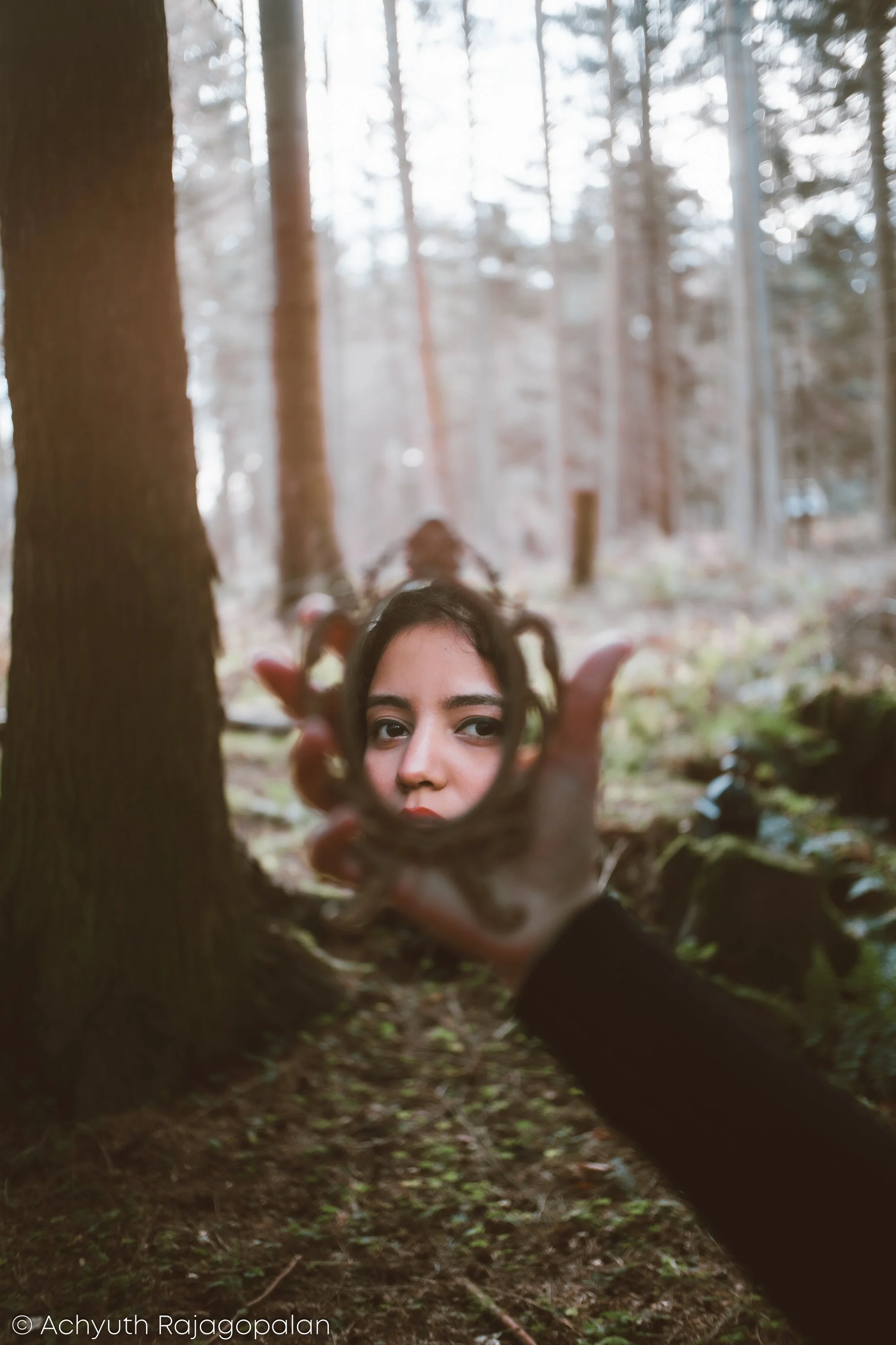 A person holding a mirror reflecting their face in a forest with tall trees and sunlight filtering through the branches.