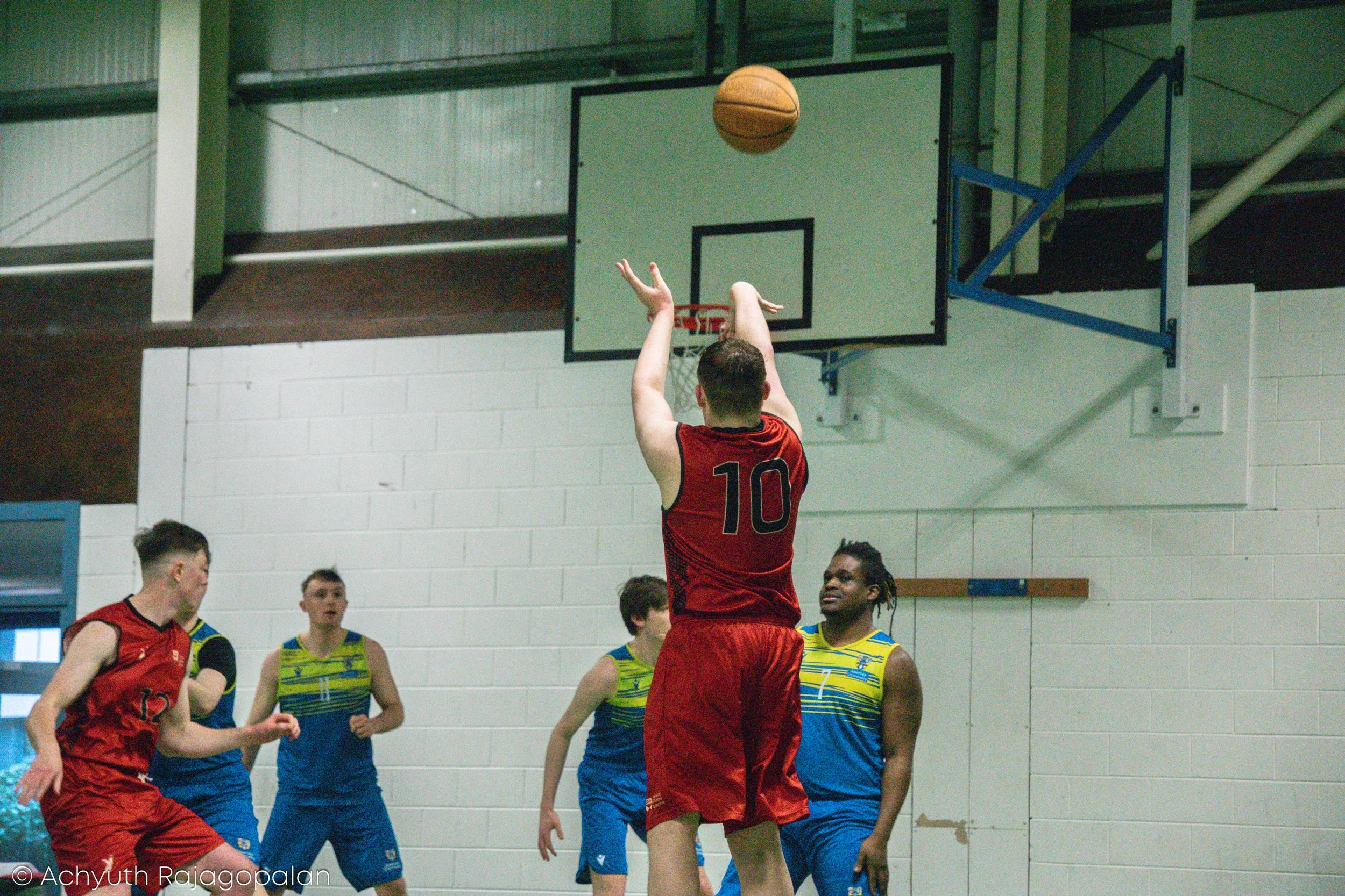 A basketball game with a player in a red jersey shooting the ball towards the hoop while other players watch nearby.