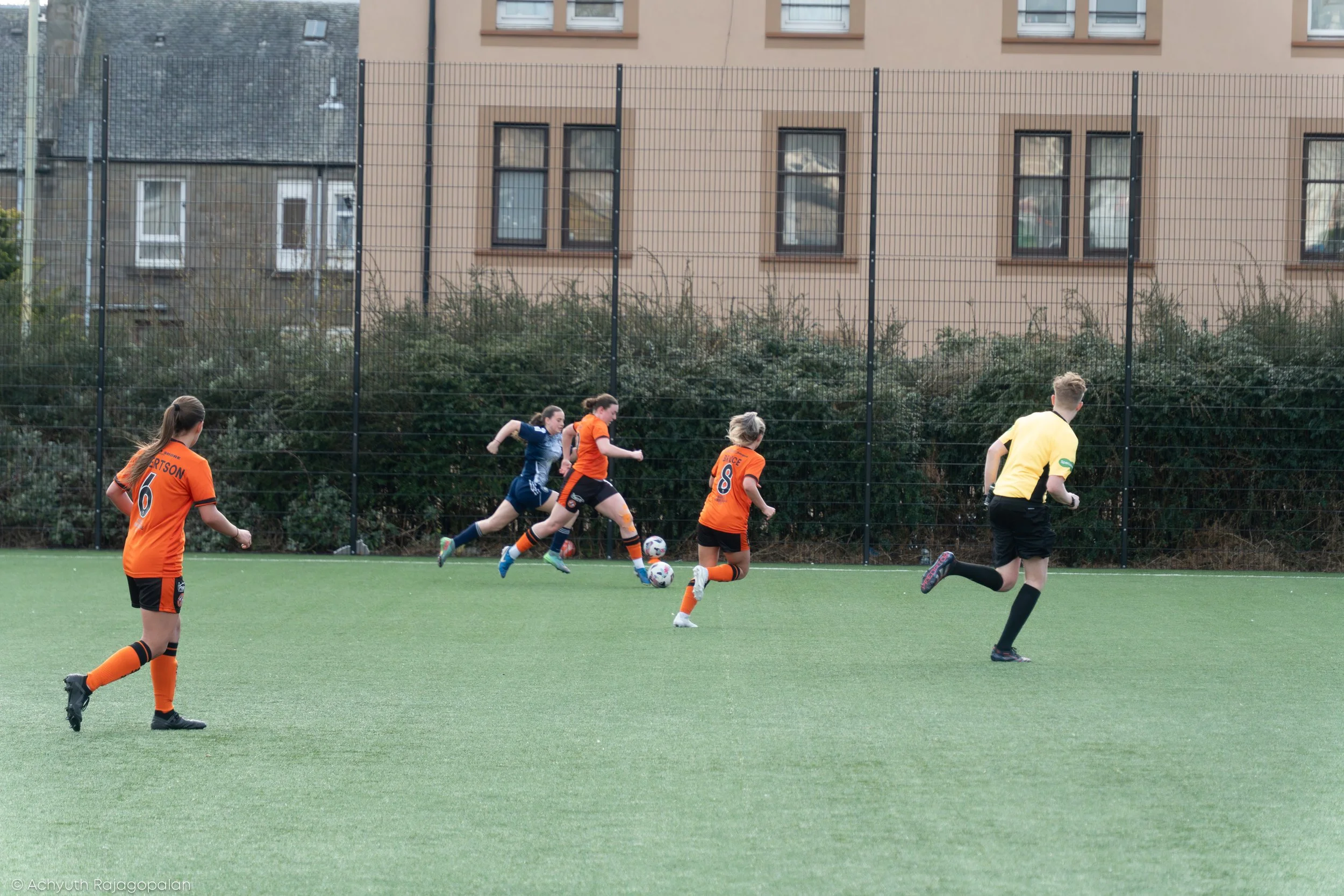 Children playing soccer on artificial turf field, with some wearing orange jerseys and one in a yellow referee shirt, against a backdrop of residential buildings.