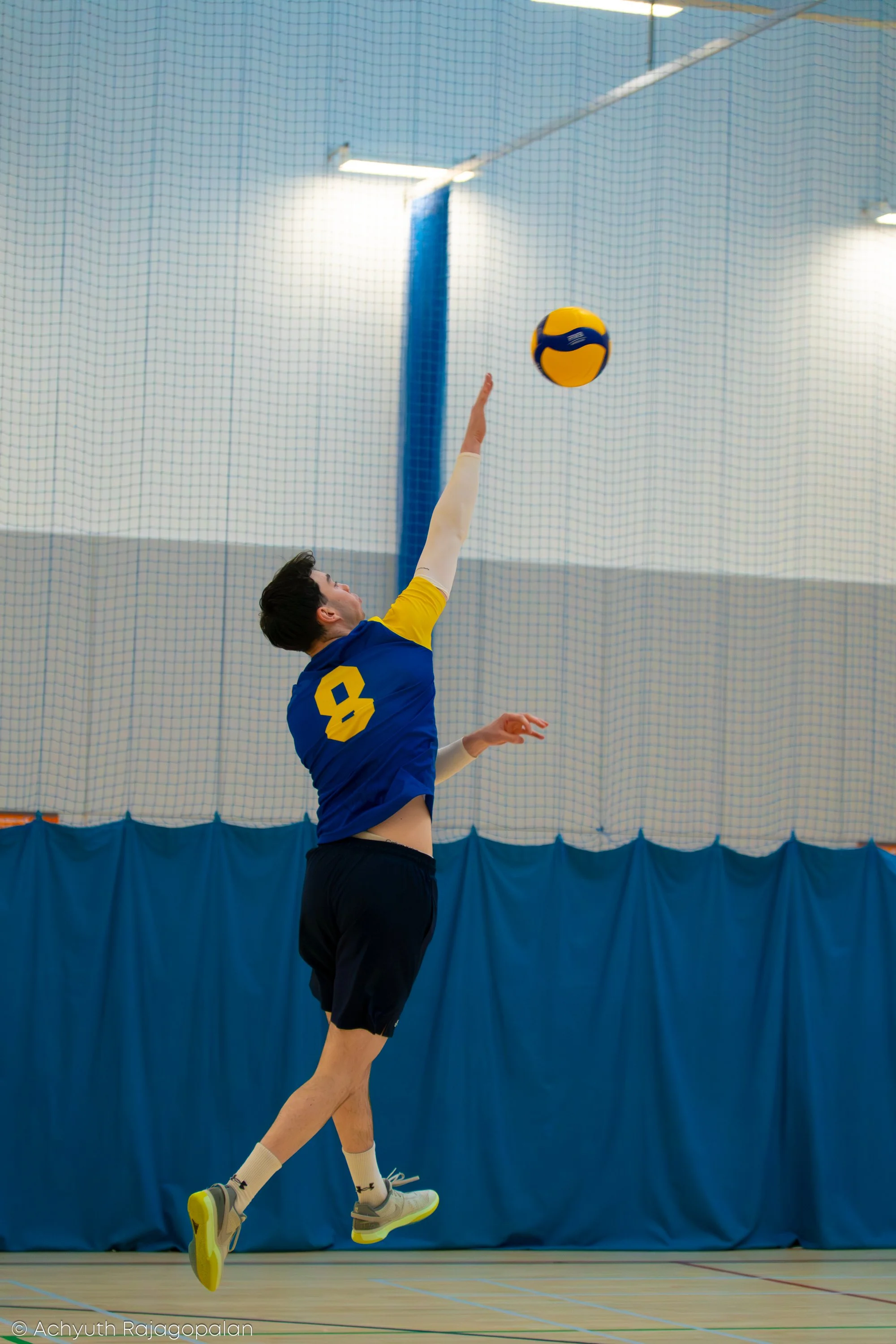 A young male volleyball player in a blue and yellow sports uniform jumping in an indoor gym, reaching to hit a volleyball over a net.