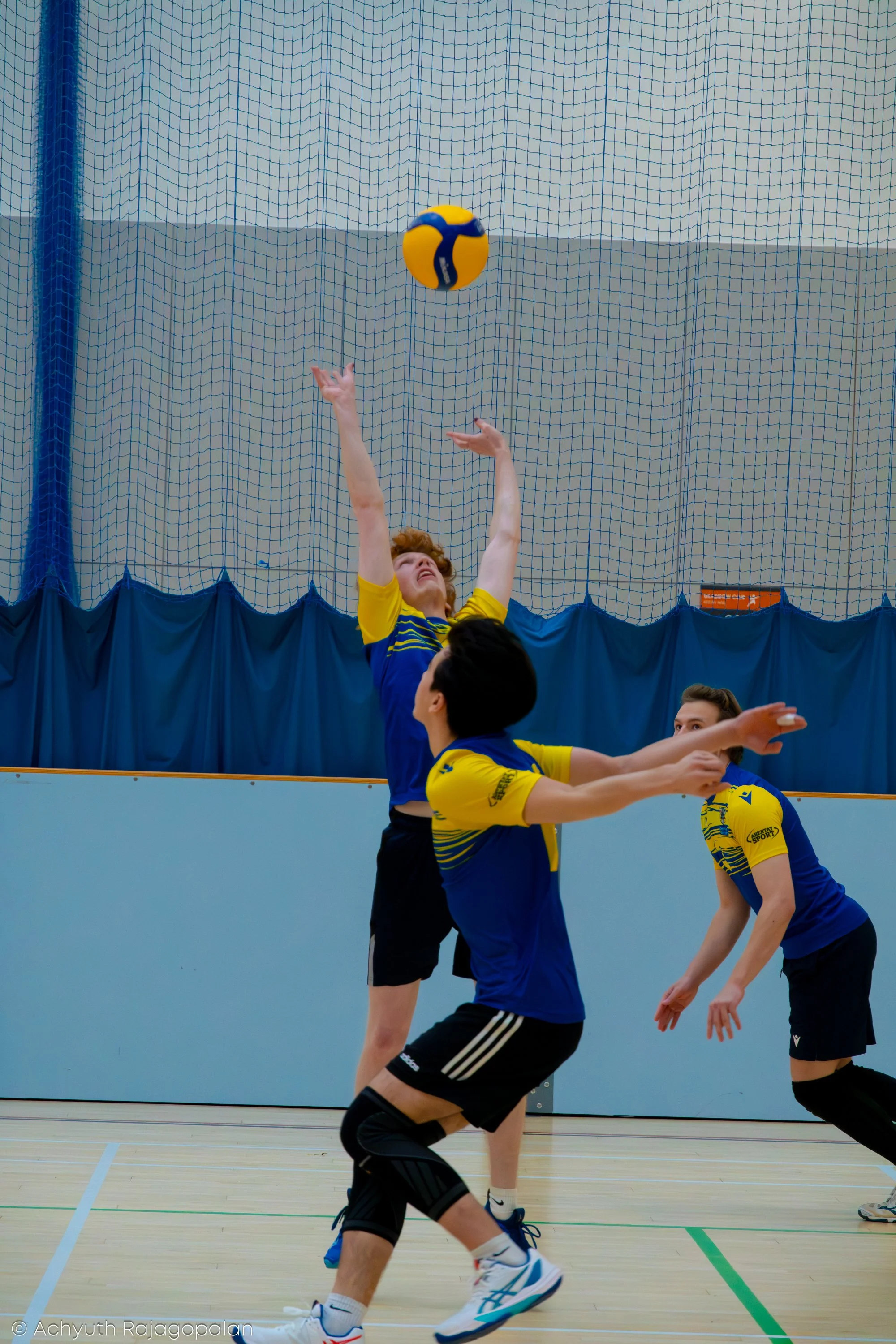 A volleyball game in progress with players jumping for the ball, which is in mid-air near the net, inside an indoor court.