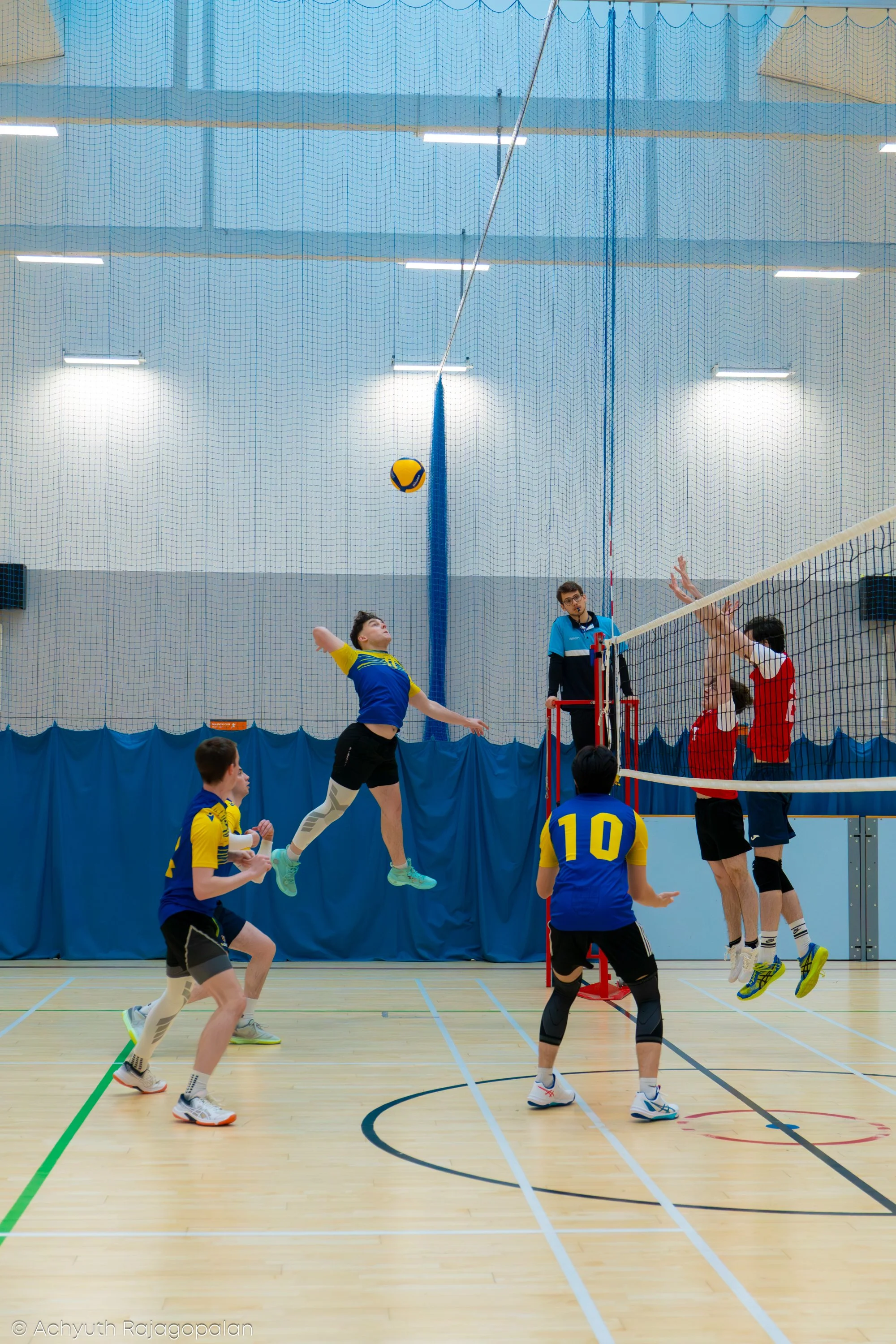 Indoor volleyball game in progress with players jumping to hit the ball over the net. The court has a light wooden floor, and spectators are not visible.