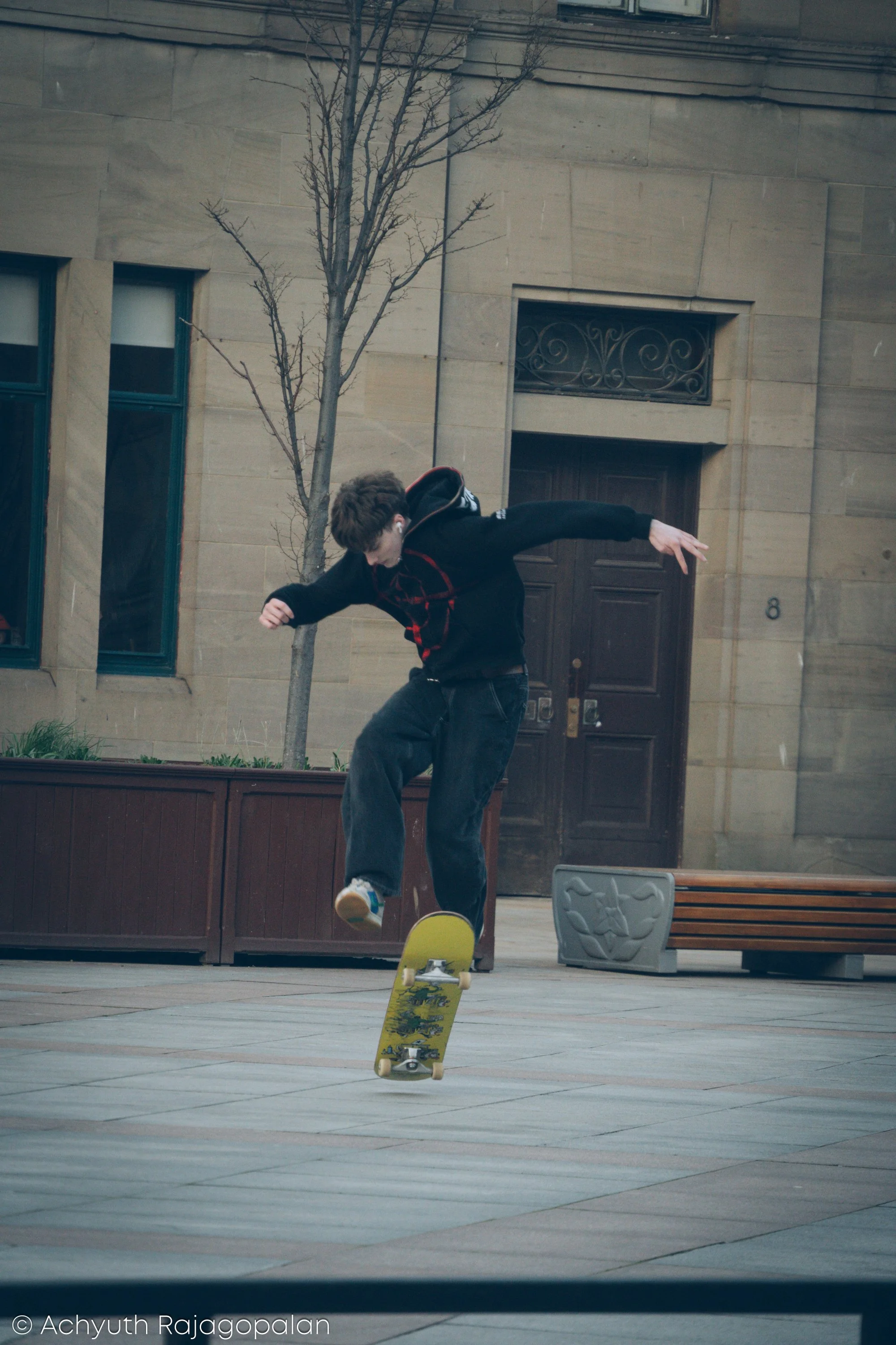 Young person skateboarding on a yellow skateboard in an outdoor urban setting, with a building and leafless tree in the background.