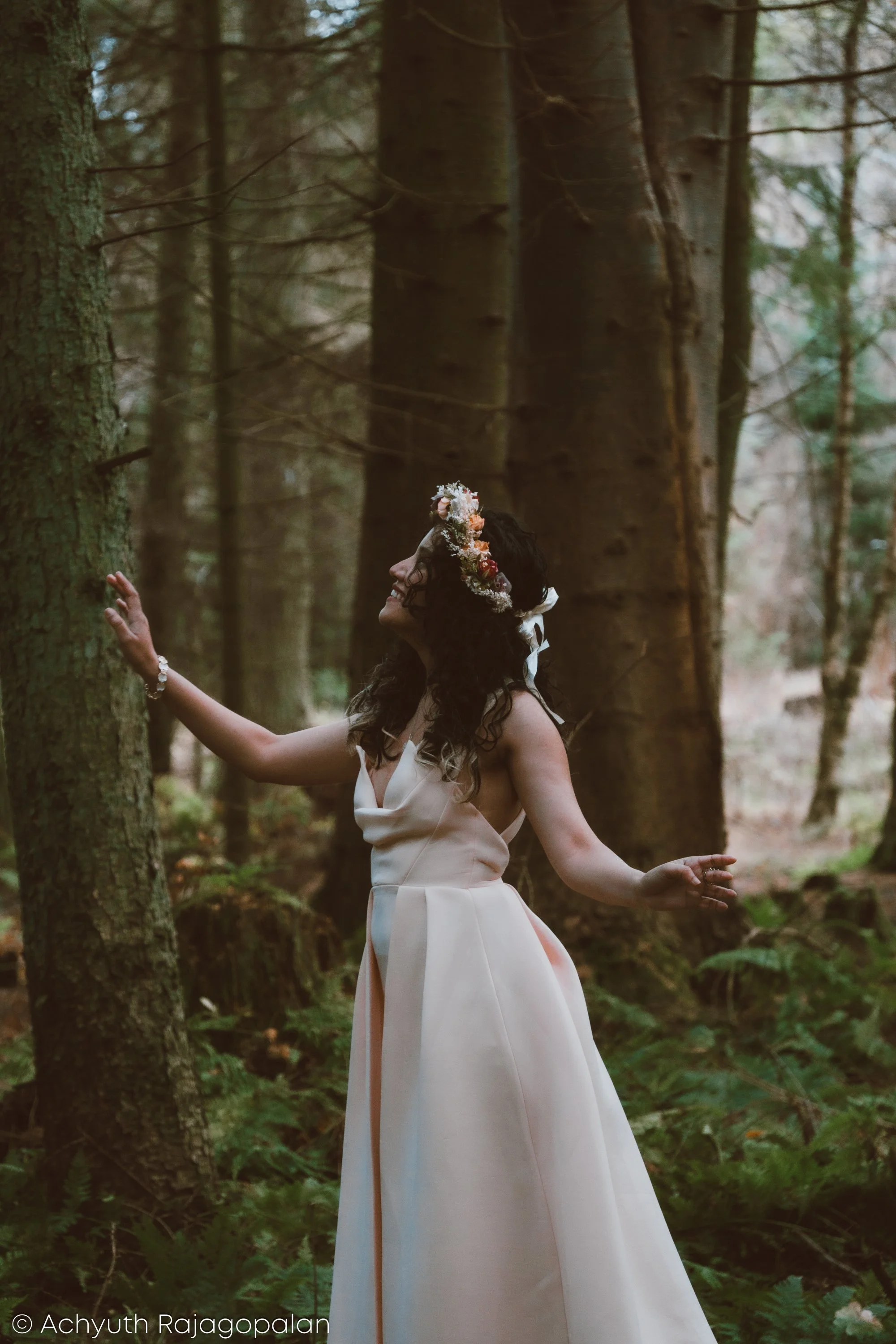 A woman in a white dress and floral crown standing in a forest with her eyes closed and arms outstretched.