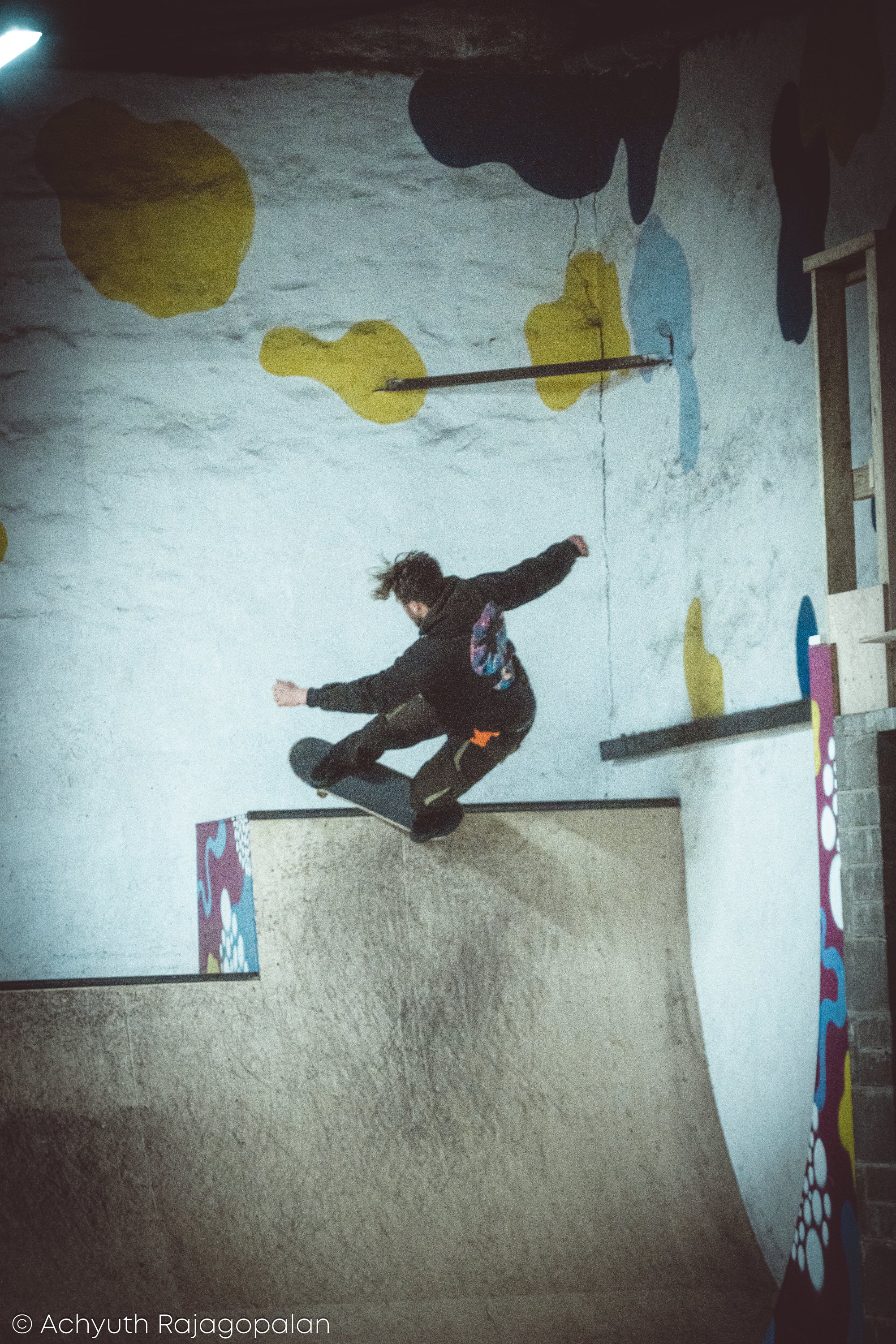 A person with curly hair in black clothing appears to be in mid-air while skateboarding indoors on a textured floor with colorful abstract wall decorations.