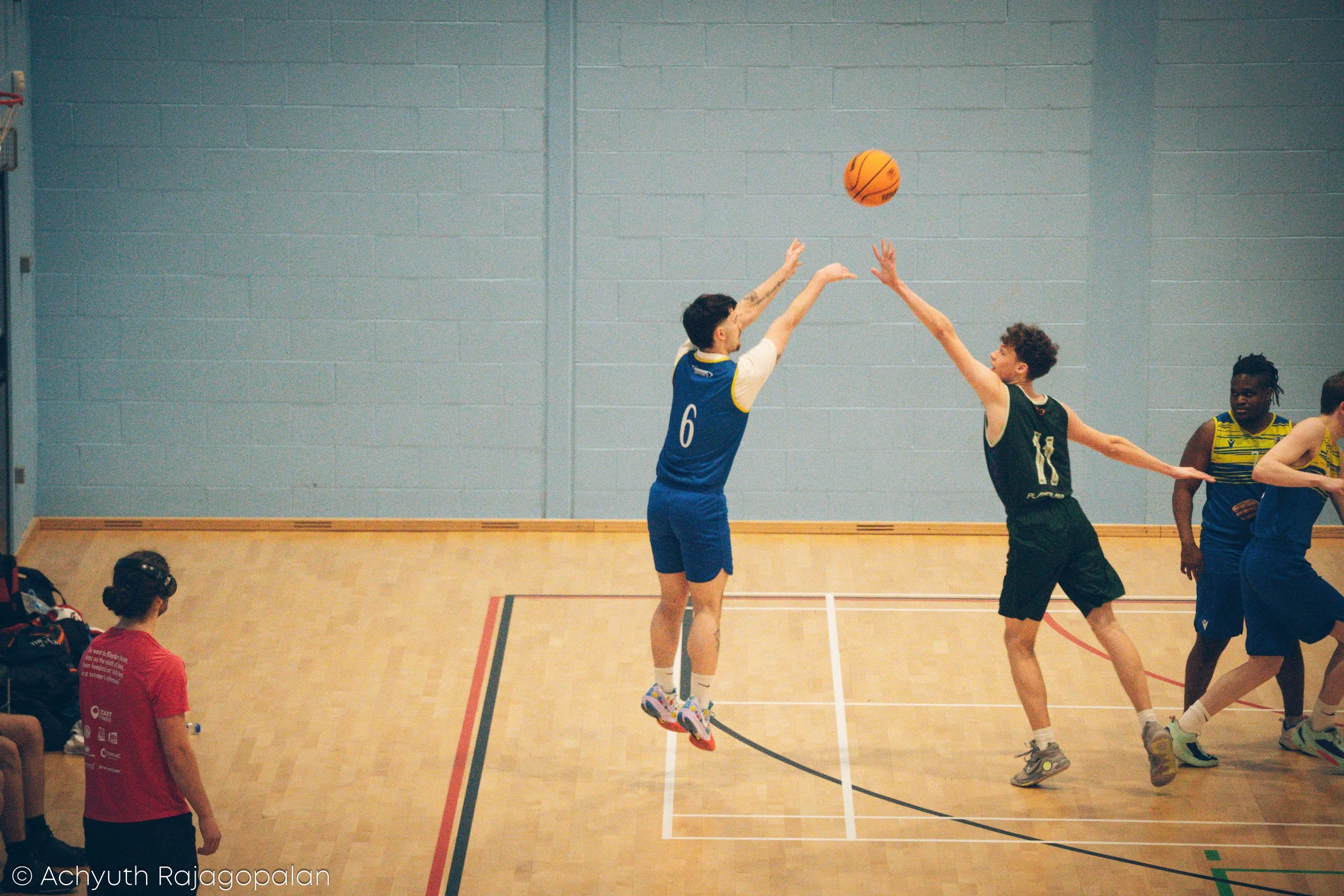 Two male basketball players are jumping for a ball in an indoor court, one wearing a blue jersey with the number 6, and the other in a black jersey with the number 10. Two other players in yellow jerseys are on the court, and a person in a red shirt 