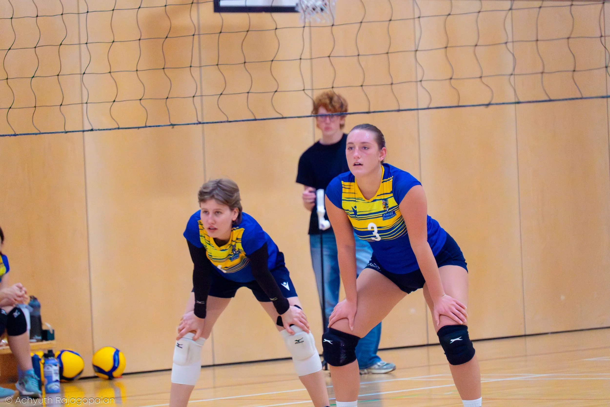 Two female volleyball players in blue and yellow uniforms are in a ready position on an indoor volleyball court, with a woman in black standing in the background behind a volleyball net.