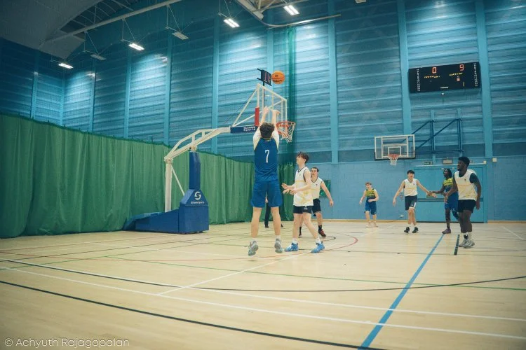 Young boys playing basketball in an indoor gym, with one player jumping to shoot the ball at the hoop.