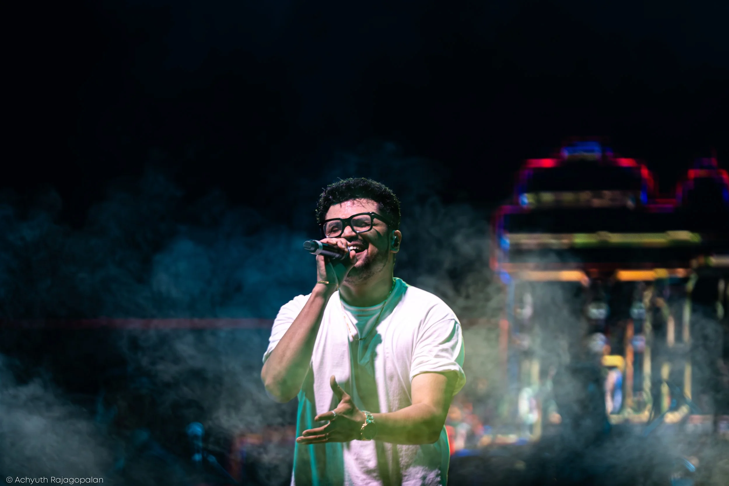 A young man with dark hair, glasses, and a white t-shirt is singing into a microphone at an outdoor event at night, with a dark background and some colorful lights.