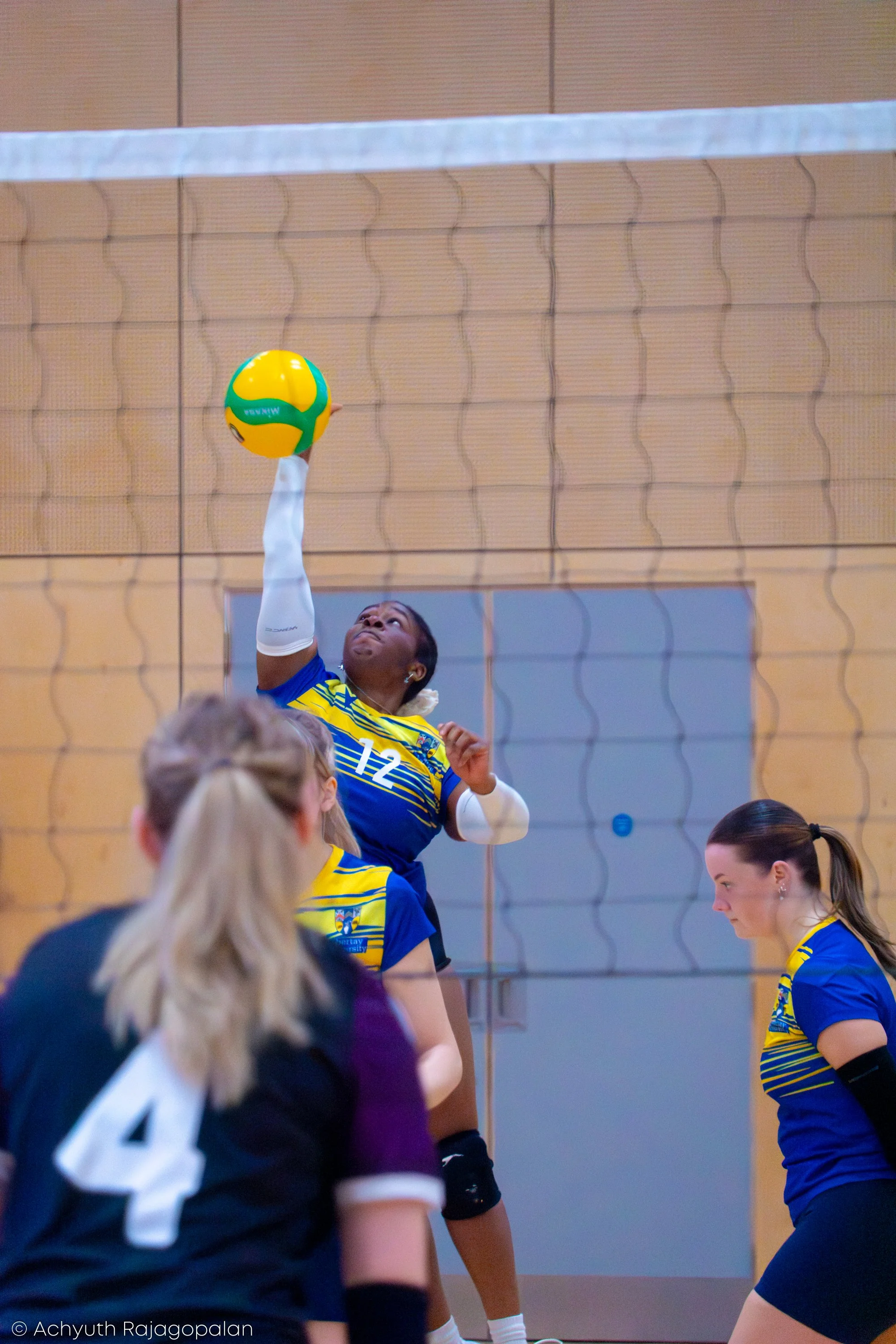 A female volleyball player in a blue and yellow uniform jumps to hit the ball during a match inside a gymnasium with a wooden wall and tennis net.