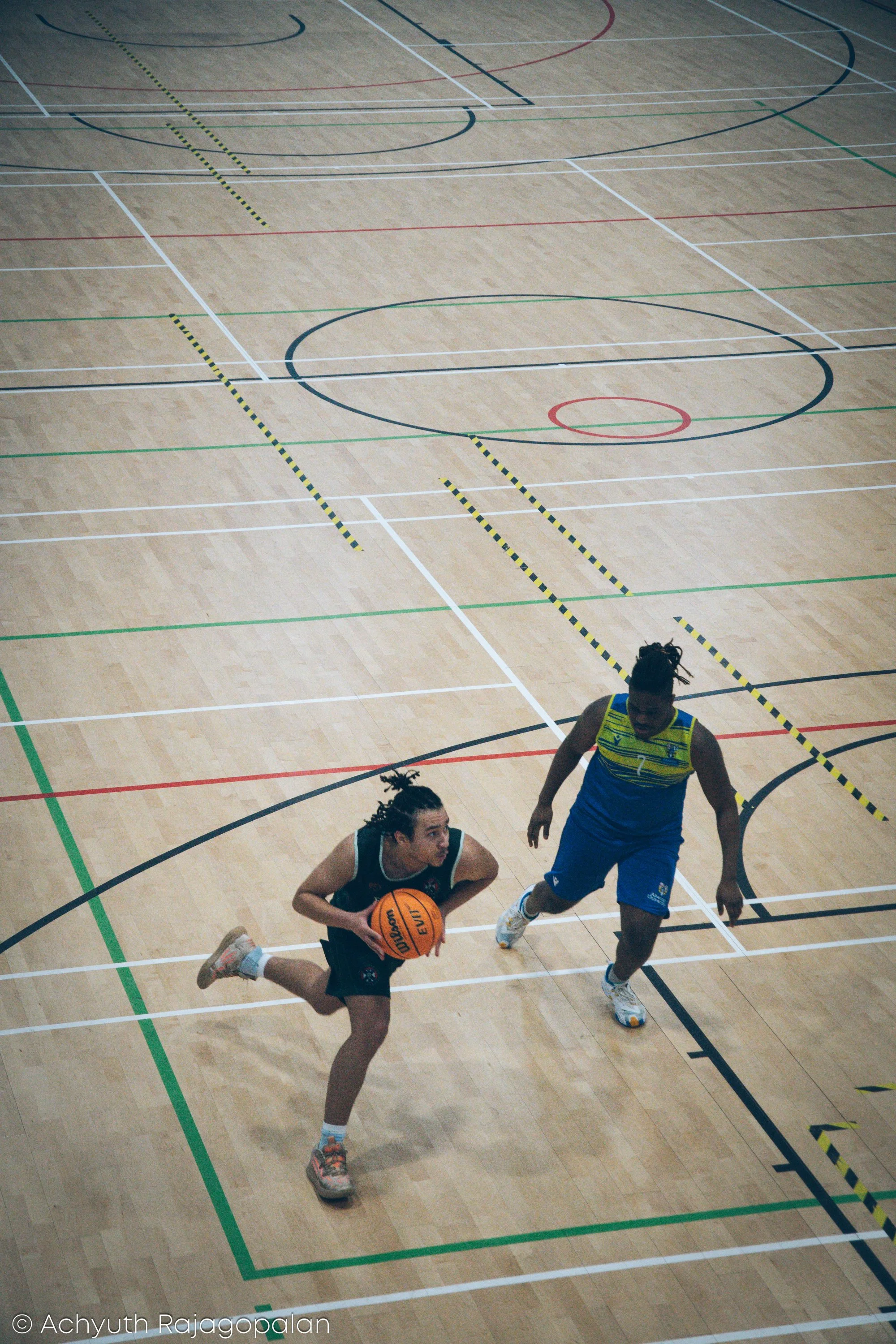 An indoor basketball court with two young men playing basketball, one is dribbling while the other is attempting to block. The court features various colored lines for different sports.