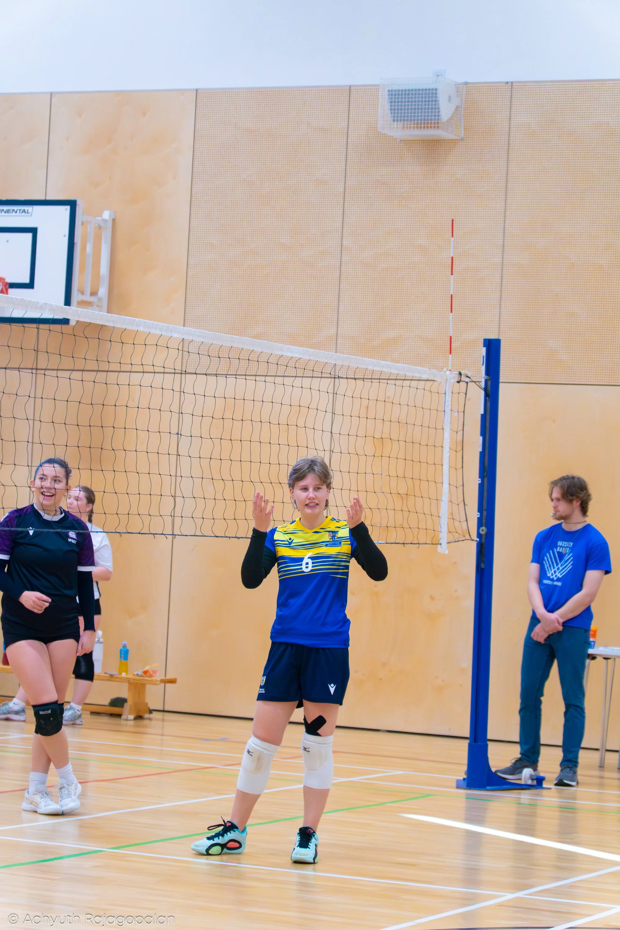 Girls playing volleyball in an indoor gym with a volleyball net and light-colored wooden walls.