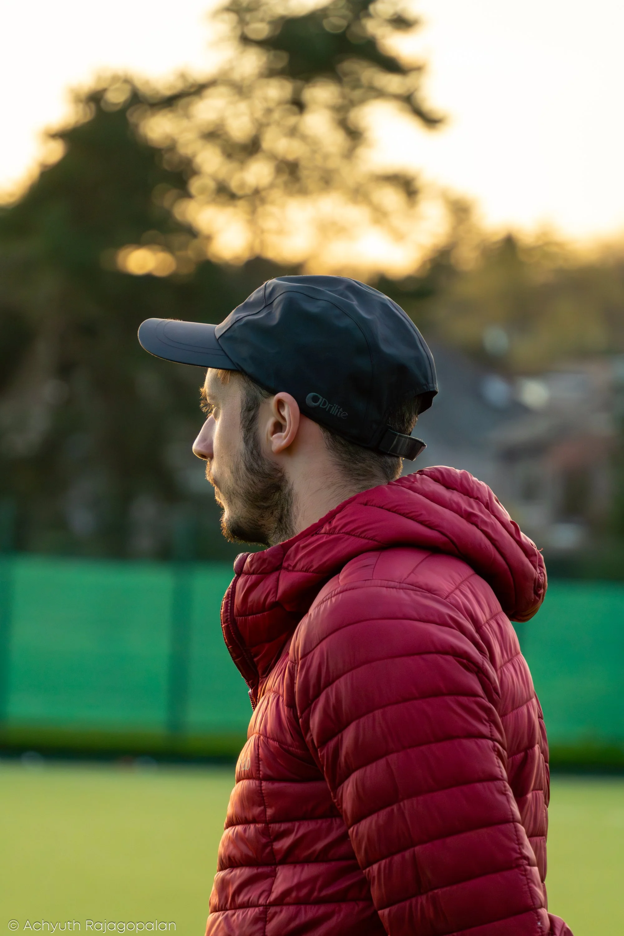 Profile of a man wearing a dark baseball cap and a red puffer jacket, standing outdoors during sunset with trees and a green field in the background.