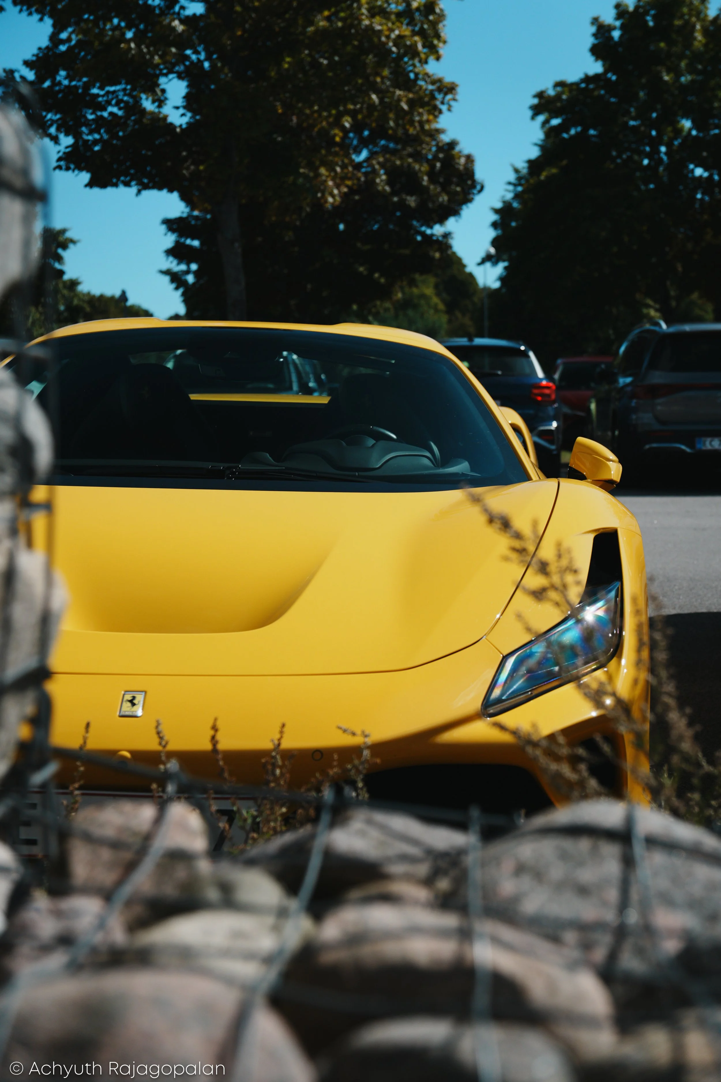 Yellow sports car parked in a lot with trees in the background, viewed through a chain-link fence.
