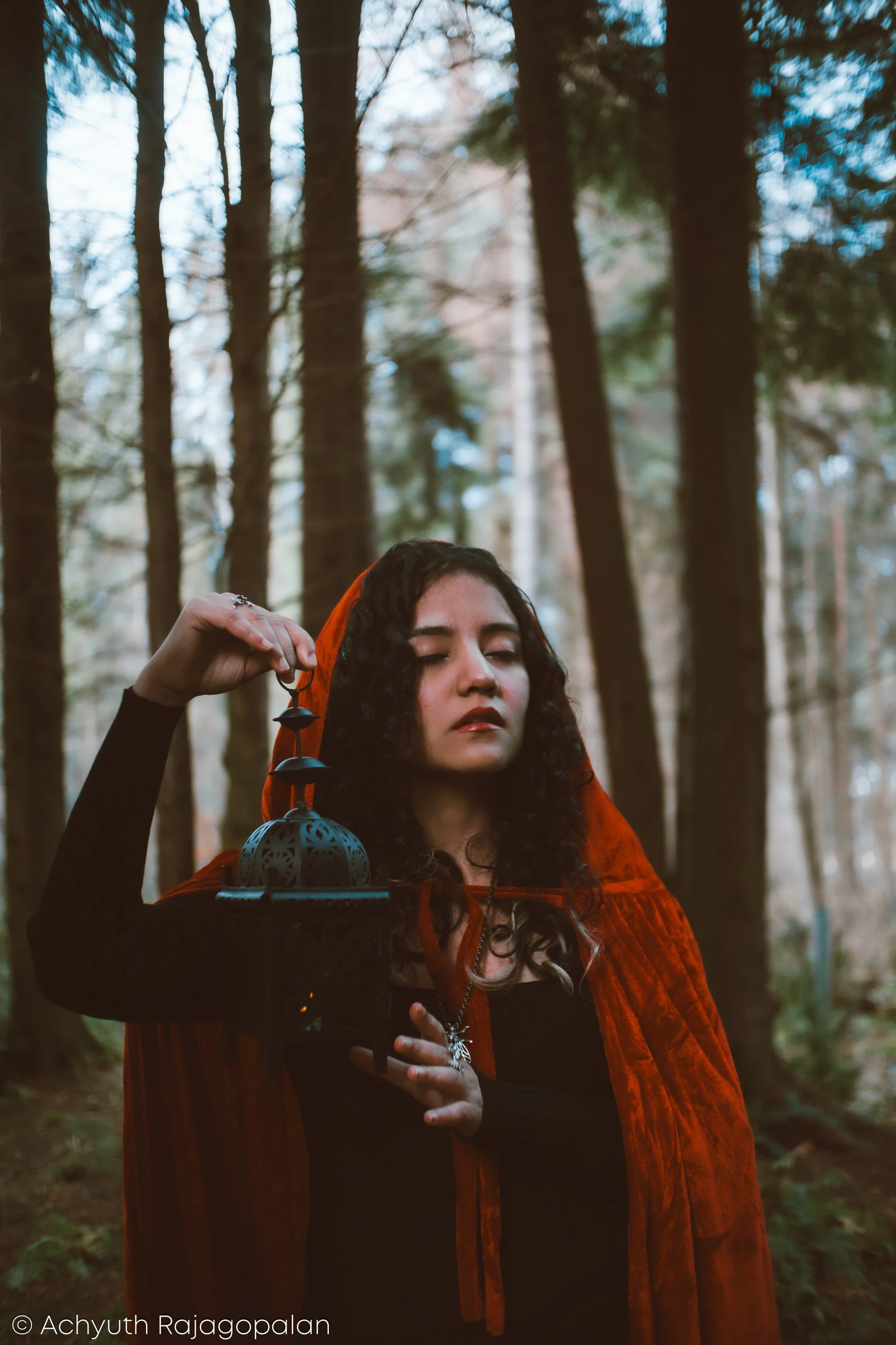 A woman with dark curly hair, wearing a red cloak and black top, standing in a forest with tall trees. She is holding a black lantern in her right hand and has her eyes closed.