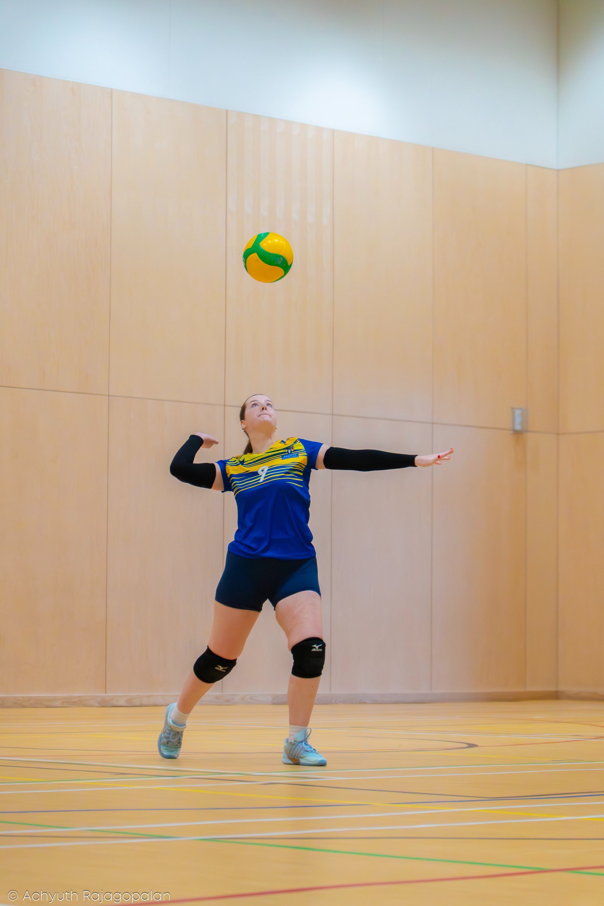 A female volleyball player in blue sports attire positioned on an indoor court, preparing to hit a green and yellow volleyball.