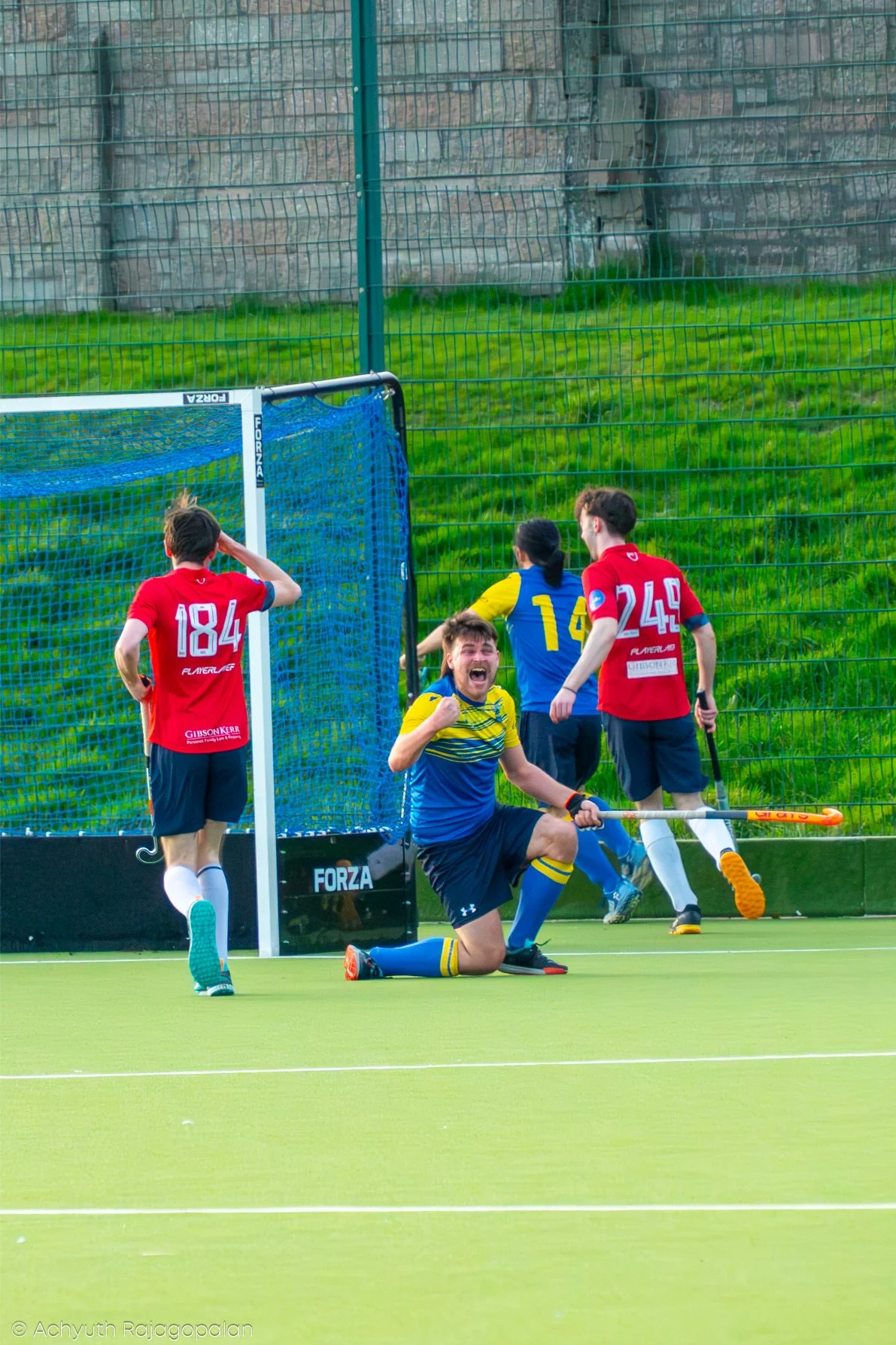 A group of field hockey players celebrating on the field after a goal, with one kneeling with a triumphant expression, others standing near the goalpost, and a green field and fence in the background.