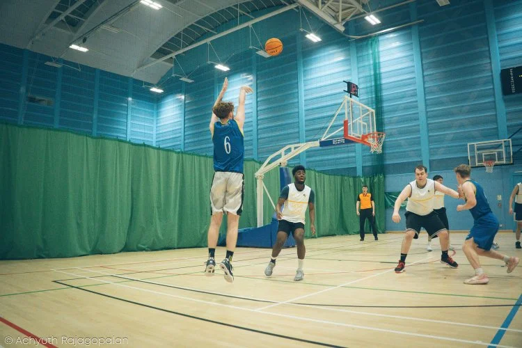 A basketball game in progress with a player in a blue jersey jumping to shoot, other players on the court, and a referee observing in an indoor gym.