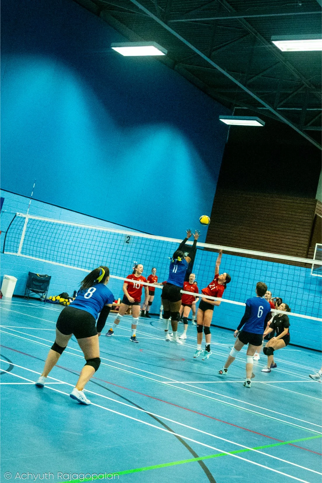 Women playing volleyball indoors, with some jumping to hit the ball over the net, others preparing to block or receive, on a blue court with a high ceiling.