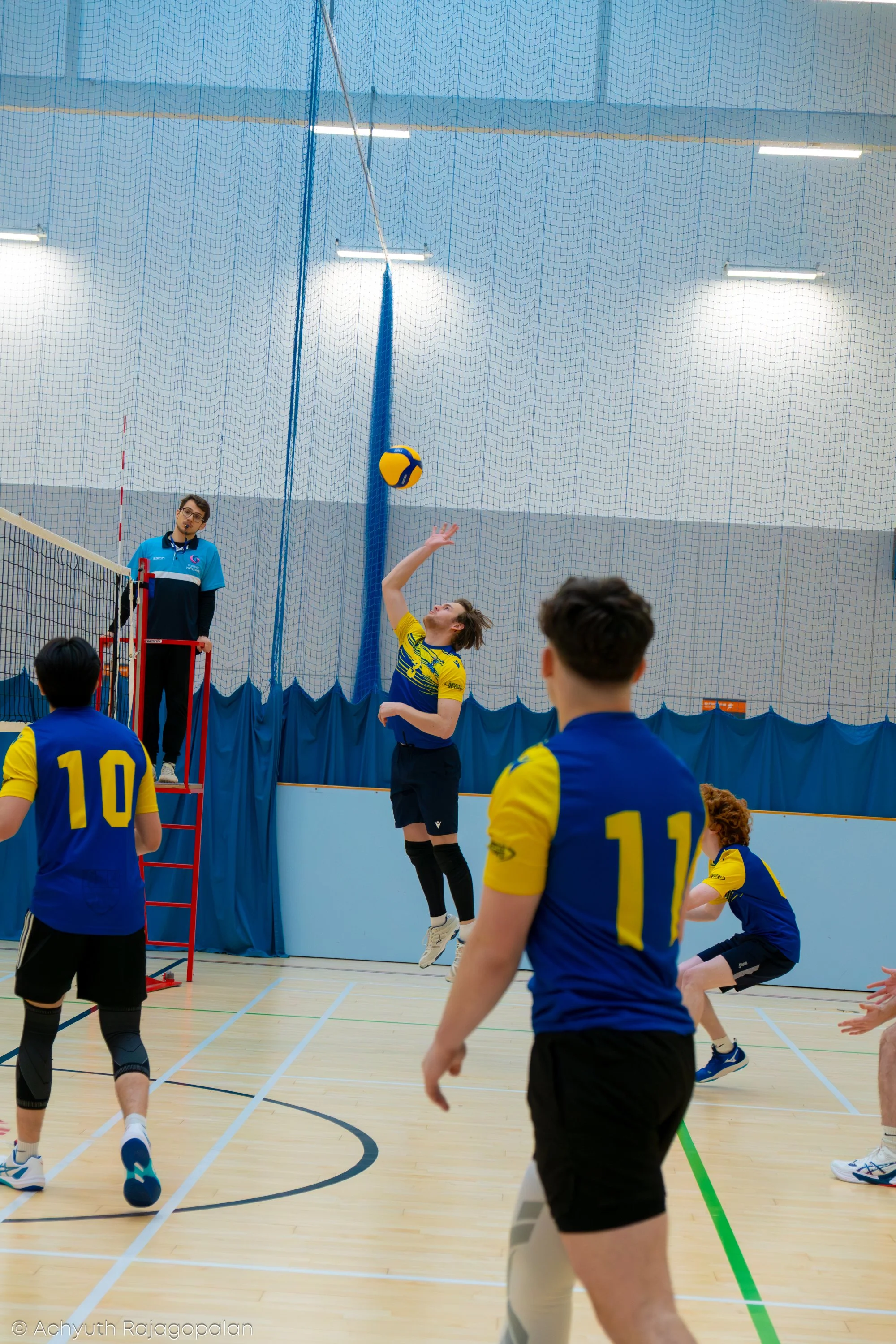 Indoor volleyball game with players in yellow and blue jerseys, one jumping to hit the volleyball, and a referee observing from the side.