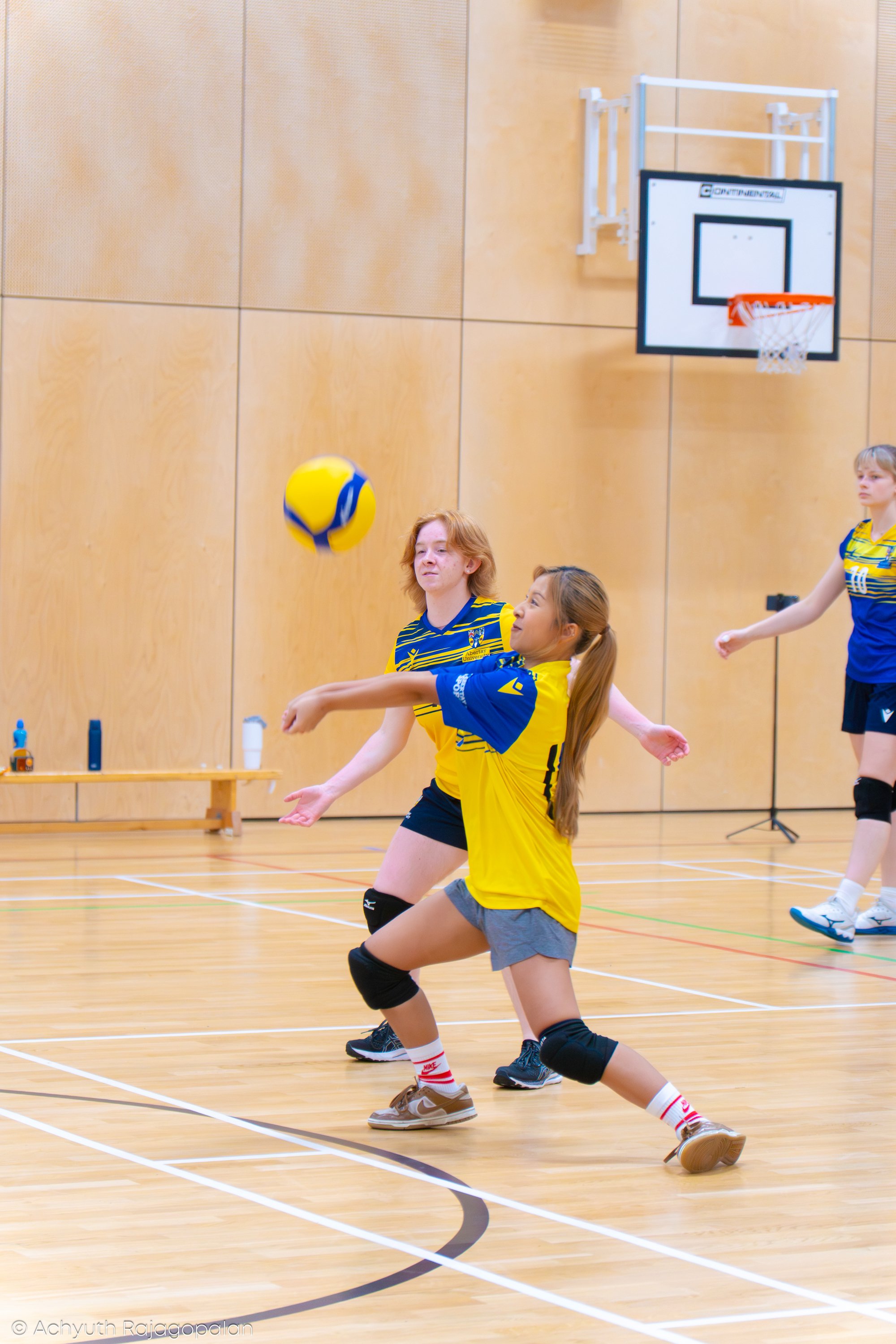 Young girls playing volleyball indoors on a gymnasium court, with one girl in a yellow and navy sports uniform reaching for the ball.