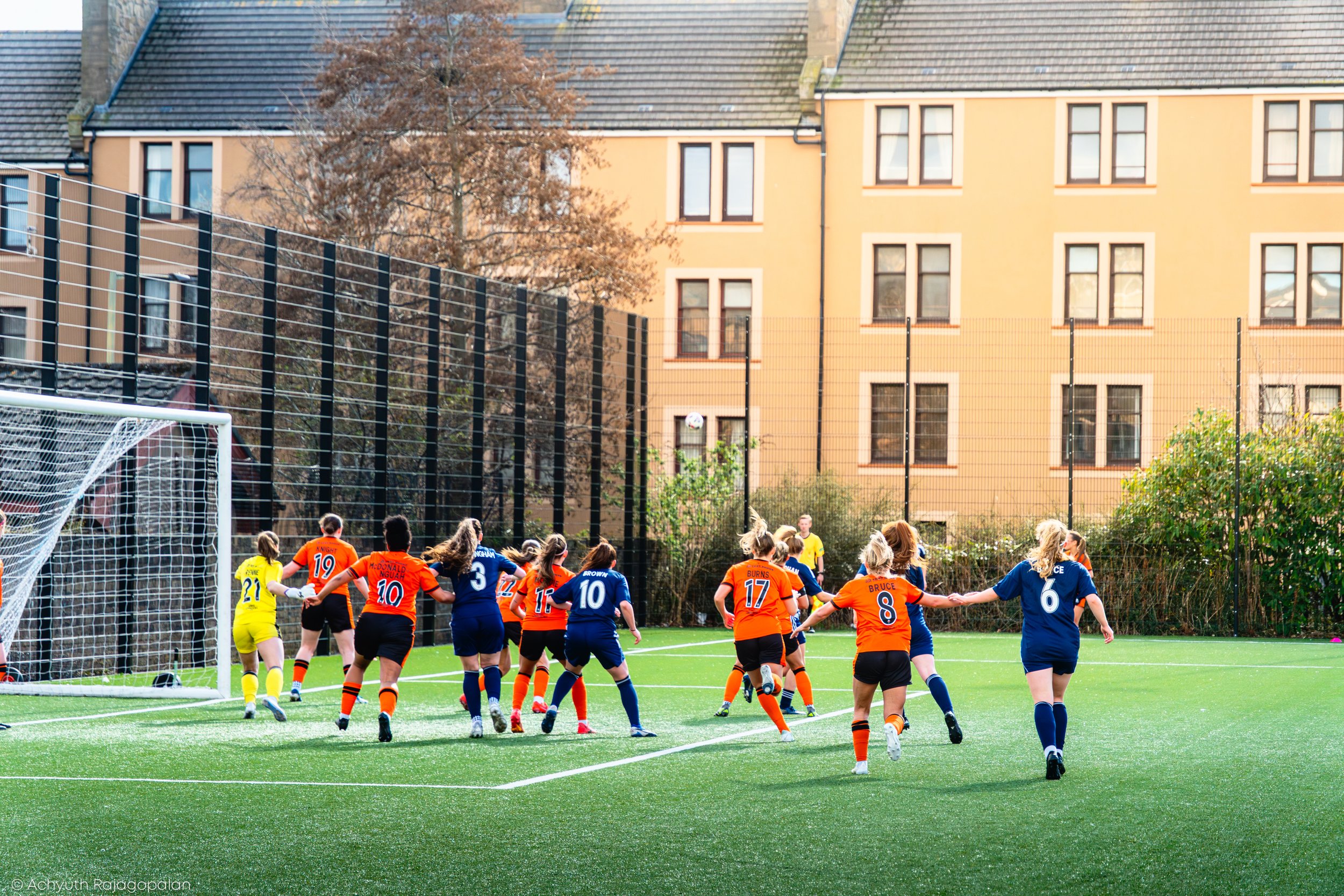 Women’s soccer game in progress with players in orange and blue jerseys on the field, near the goalpost, with residential buildings and trees in the background.