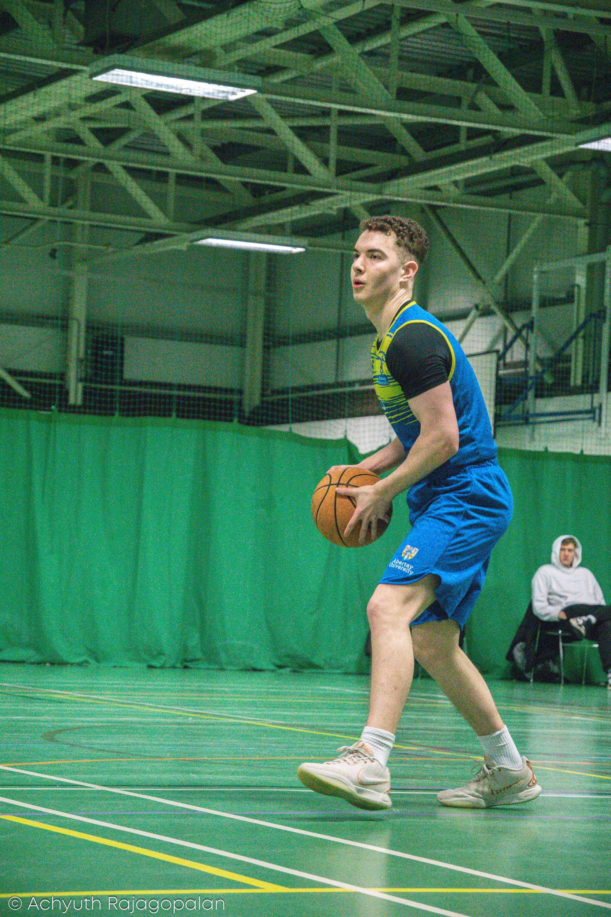 A young man playing basketball on an indoor court, wearing a blue sports uniform and white sneakers, with a woman sitting nearby watching.
