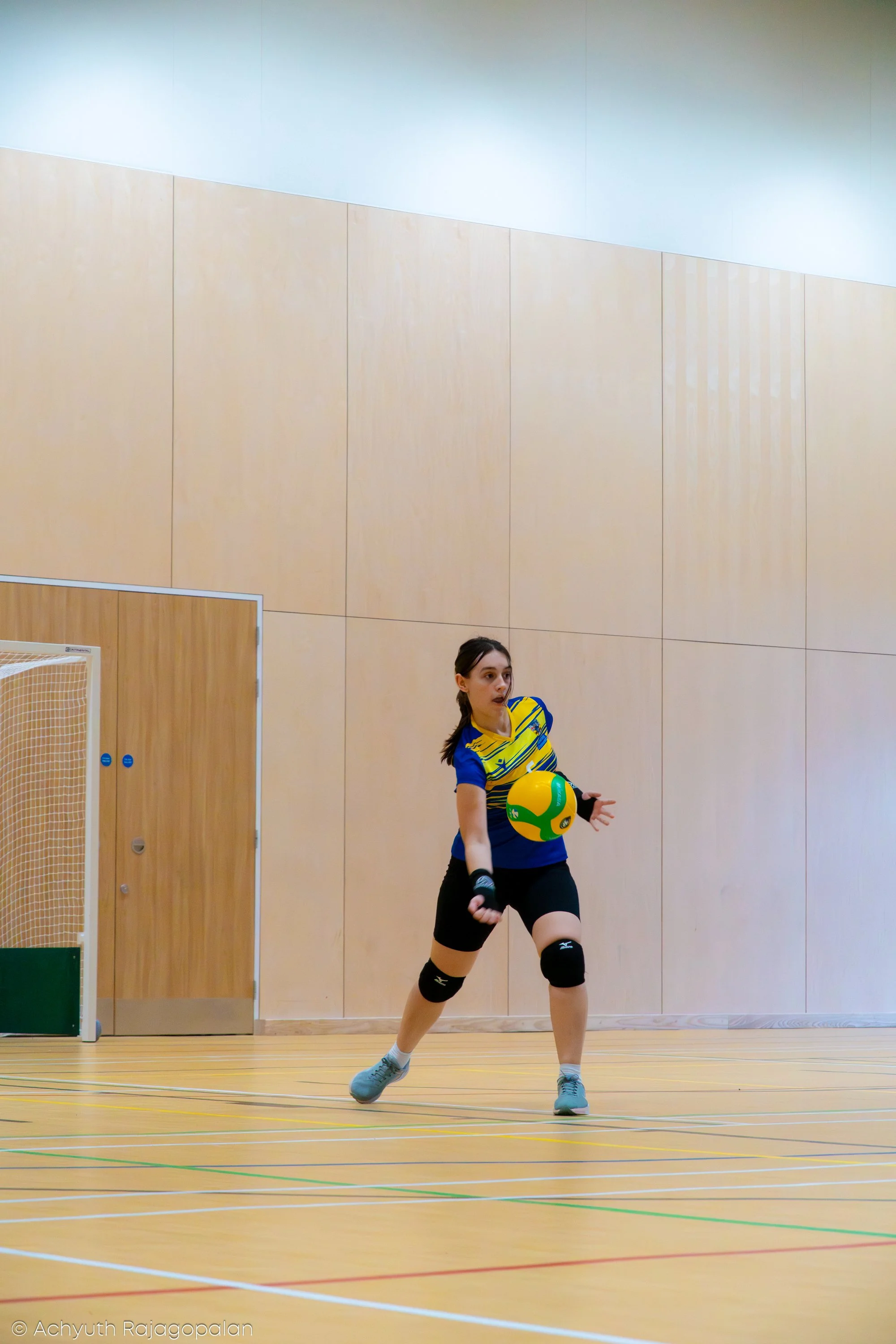 An indoor volleyball court with a female player in a blue and yellow uniform holding a green and yellow volleyball, with a neutral-colored wall and part of a volleyball net visible in the background.