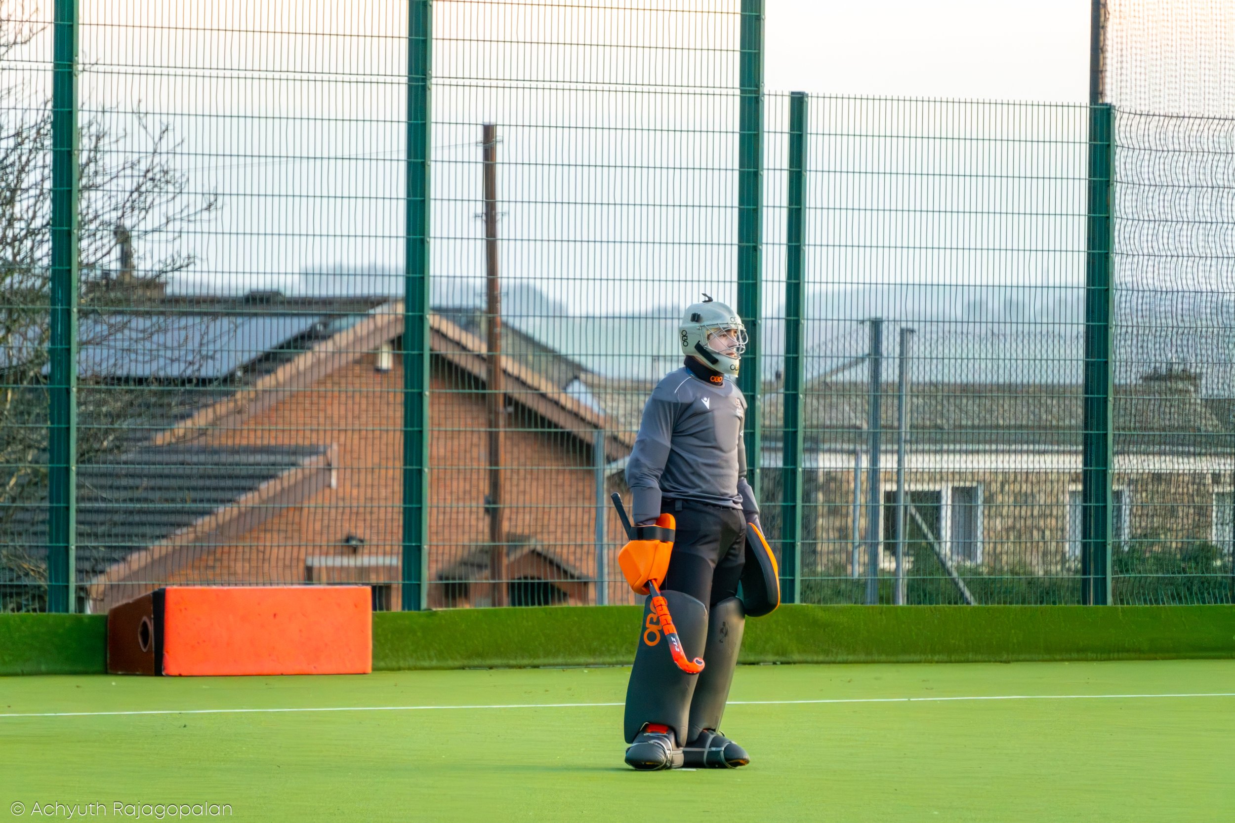 A field hockey player standing on a green field, wearing grey sports attire, a helmet, and holding a hockey stick. The player is behind a tall green fence with a house and cloudy sky in the background.