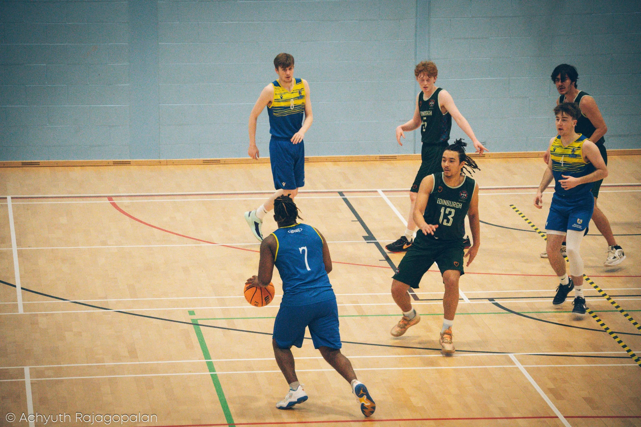 A group of young men playing basketball on an indoor court. Some are wearing black jerseys with 'Edinburgh' printed on them, and others are wearing blue and yellow jerseys. One player is dribbling the ball while others are watching or preparing to mo