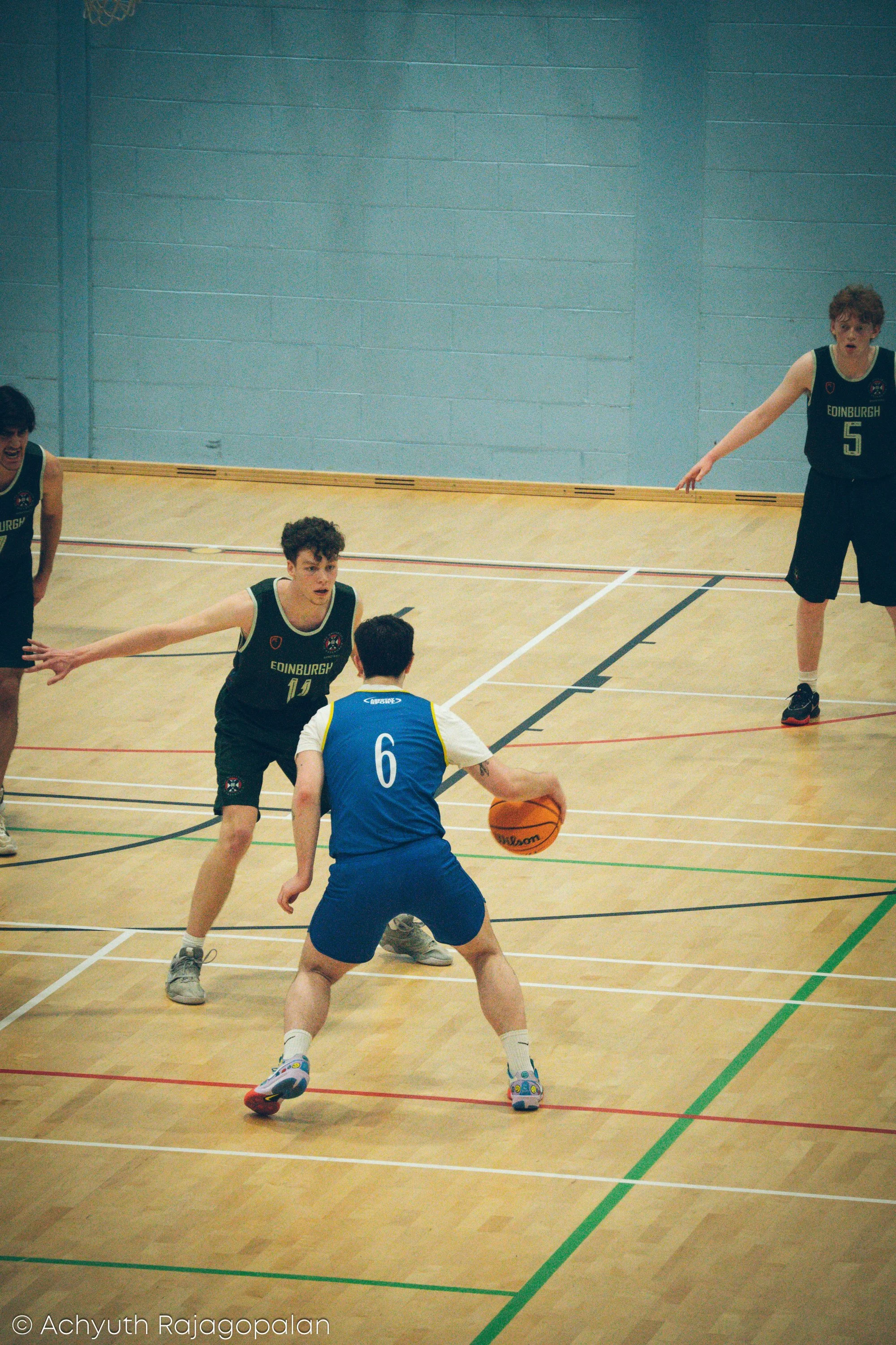 A group of young male basketball players in a gymnasium during a game, with one player dribbling the ball while others are defending.
