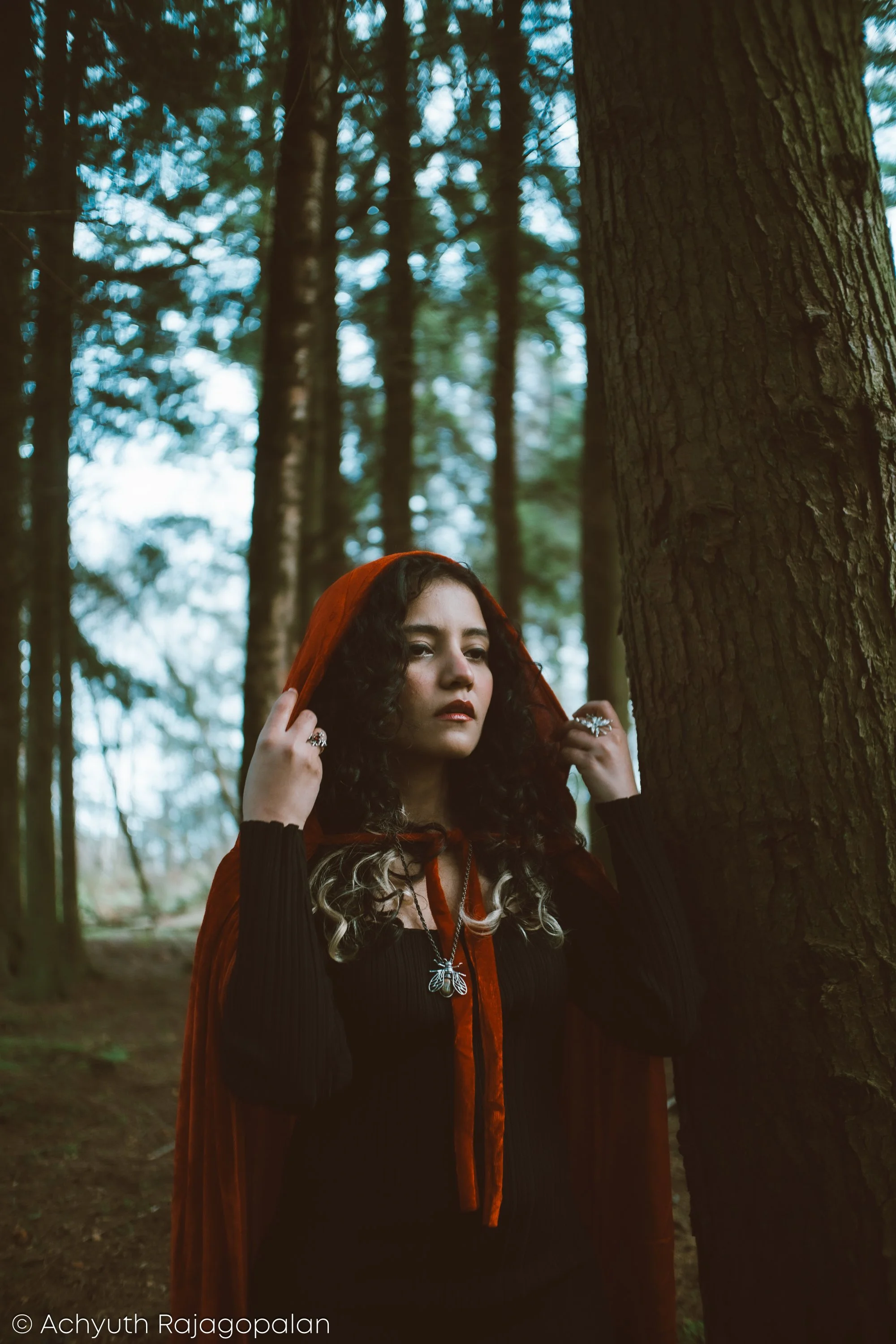 A woman with curly hair, wearing a black outfit, a necklace with a butterfly pendant, and an orange or red hooded cape, is standing in a forest next to a large tree.