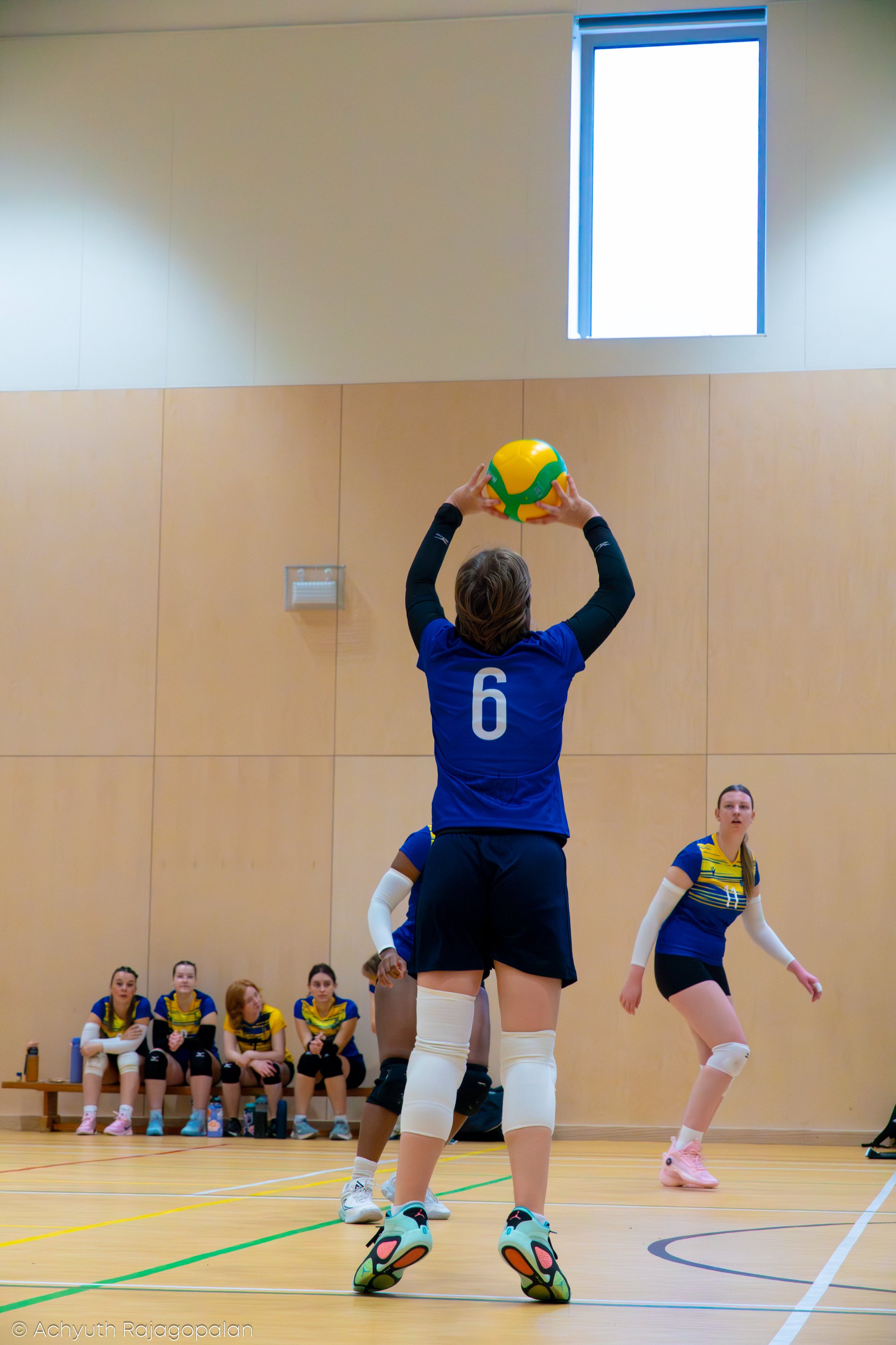 A female volleyball player in a blue jersey with the number six, preparing to serve with a volleyball in an indoor gymnasium. Behind her, four teammates sit on a bench, and two other players stand on the court. The gym has a wooden floor and a high w