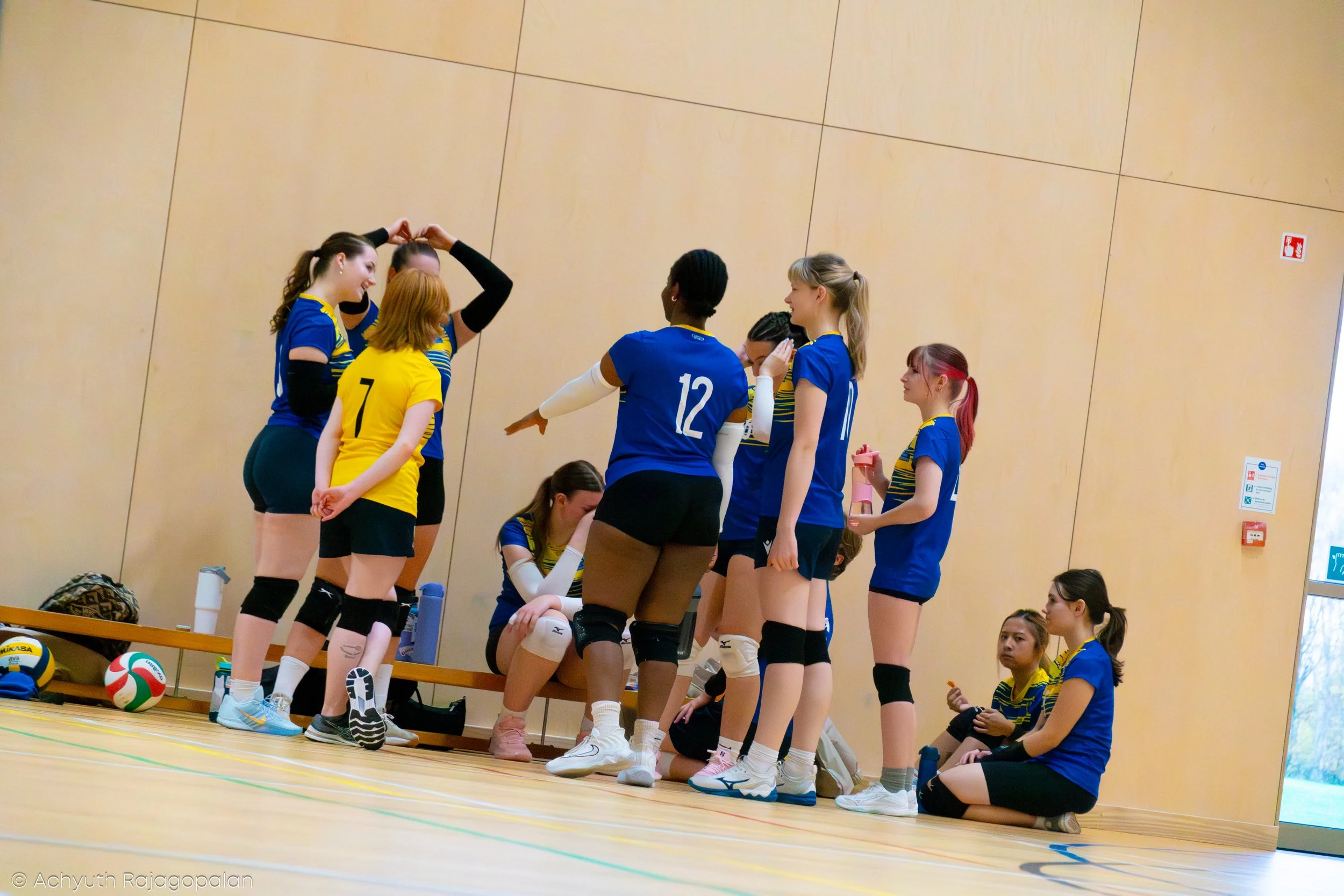 Volleyball team members in blue and yellow jerseys gather during a timeout in a gymnasium, with volleyball equipment and water bottles nearby.