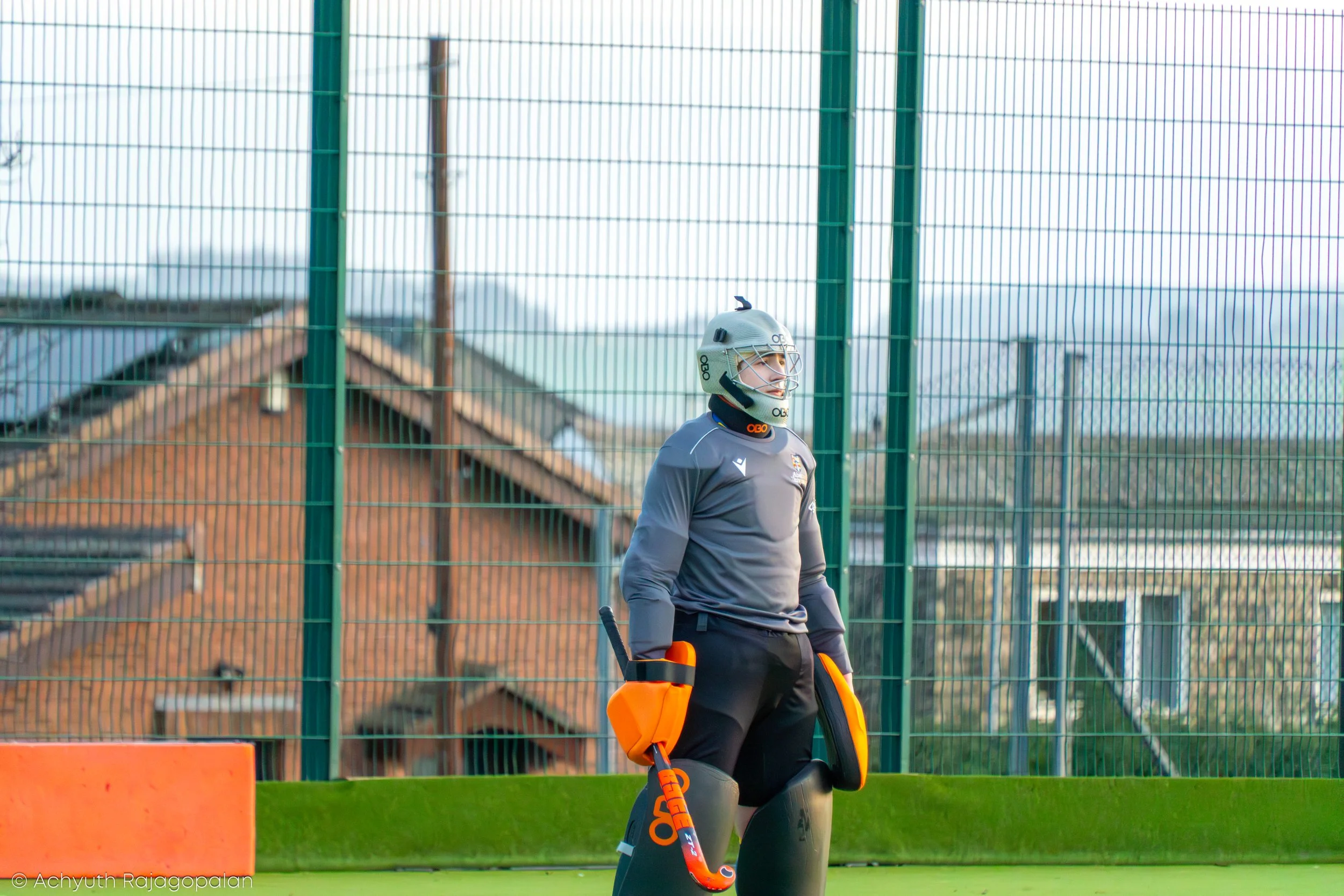 A woman dressed in sports gear, wearing a helmet and holding a hockey stick, standing outdoors near a green fence.
