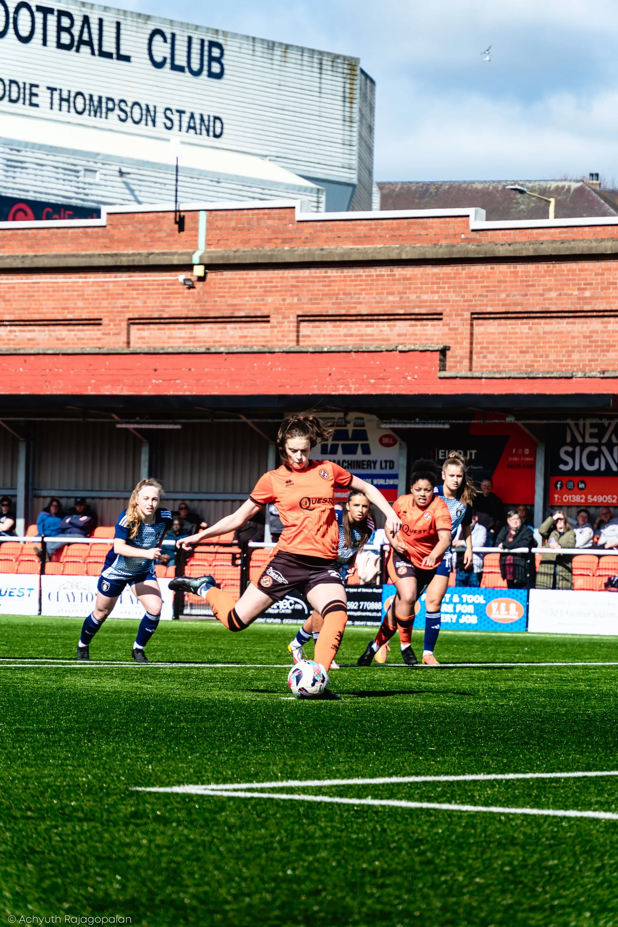 A women's soccer game with players in orange and blue uniforms on a green field, focusing on a player in orange kicking the ball. There are spectators and advertisements in the background.