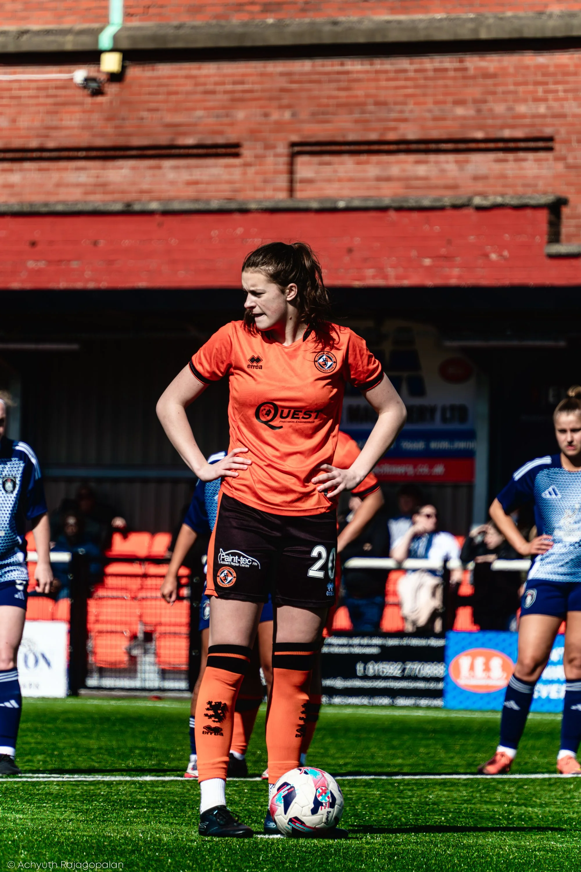 A female soccer player in an orange jersey stands with hands on hips behind a soccer ball on a green field, with other players and a brick wall in the background.