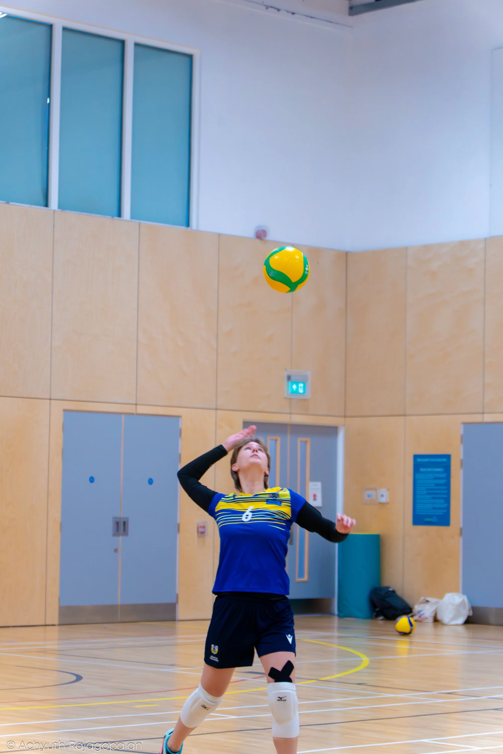 A young female volleyball player in a blue and yellow uniform is inside a gymnasium, preparing to hit a volleyball. She is wearing knee pads, white socks, and sports shoes, with her right arm raised and hand open. The gym has light-colored wood floor