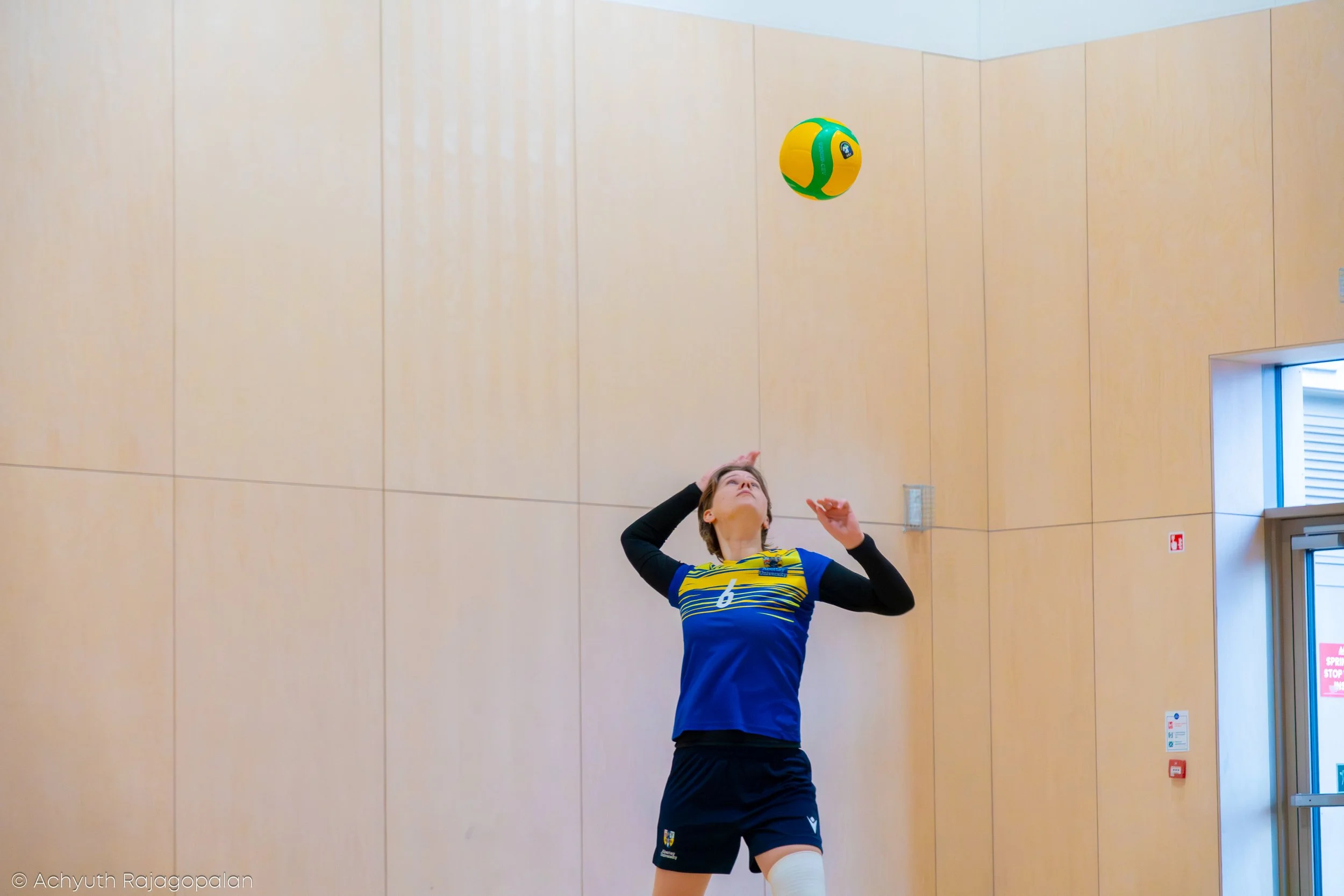 A female volleyball player wearing a blue and yellow uniform with the number six is preparing to hit a volleyball inside a gymnasium with wooden walls and a door on the right side.