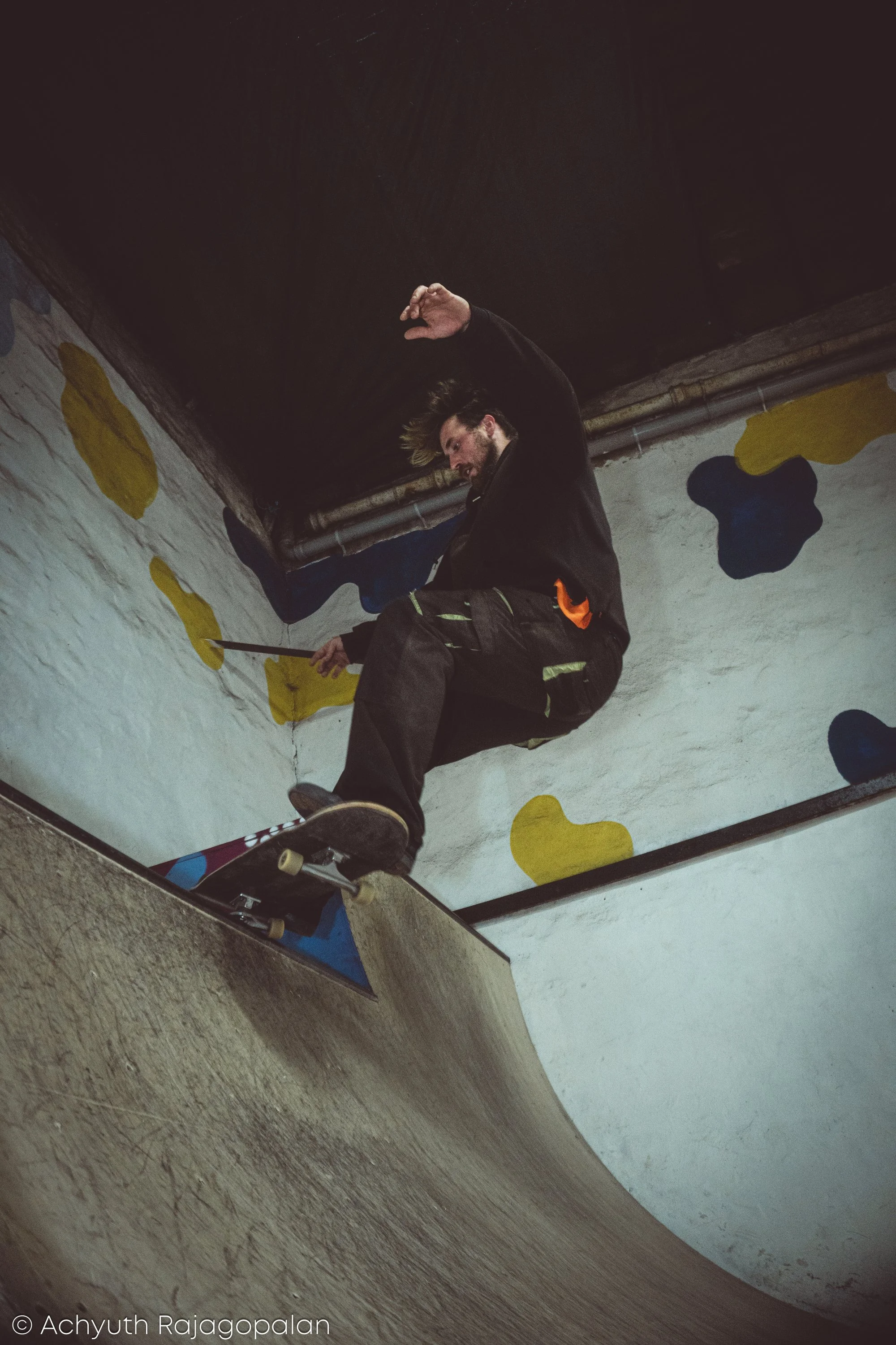 A person with beard and long hair, wearing black jacket and dark pants, sitting on skateboarding ramp surrounded by painted yellow and black abstract shapes, holding a skateboard.