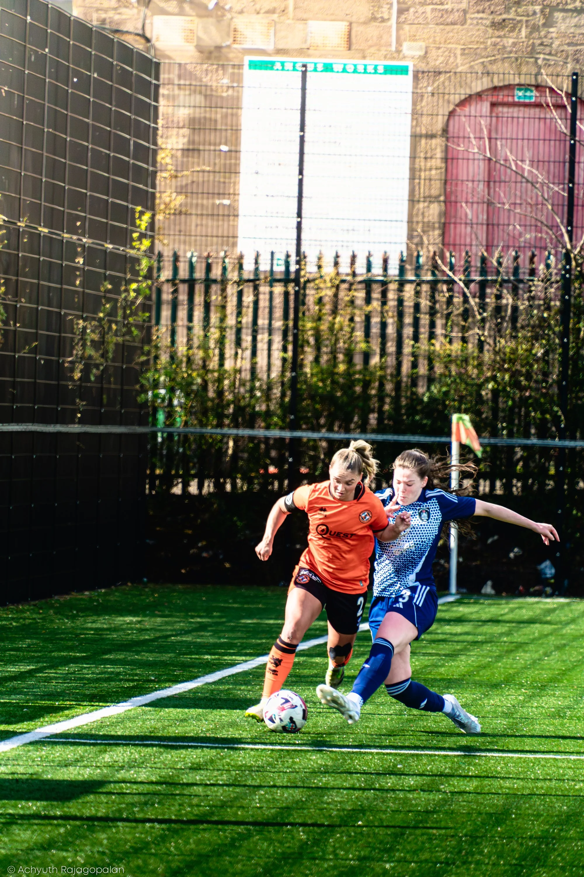 Two female soccer players competing for ball on a green field, one in an orange jersey and the other in a blue jersey, with a wall, a tree, and a gate in the background.
