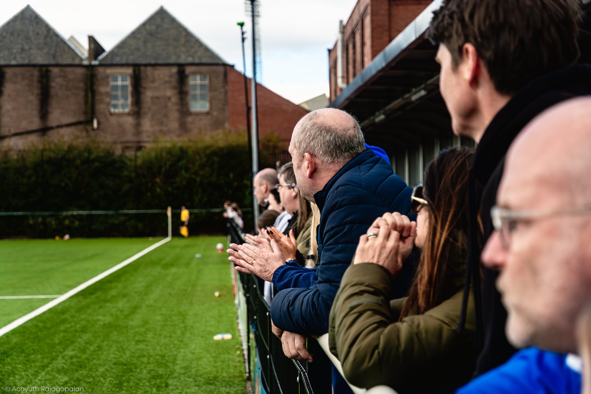 People sitting on a bench by a soccer field, watching a game.